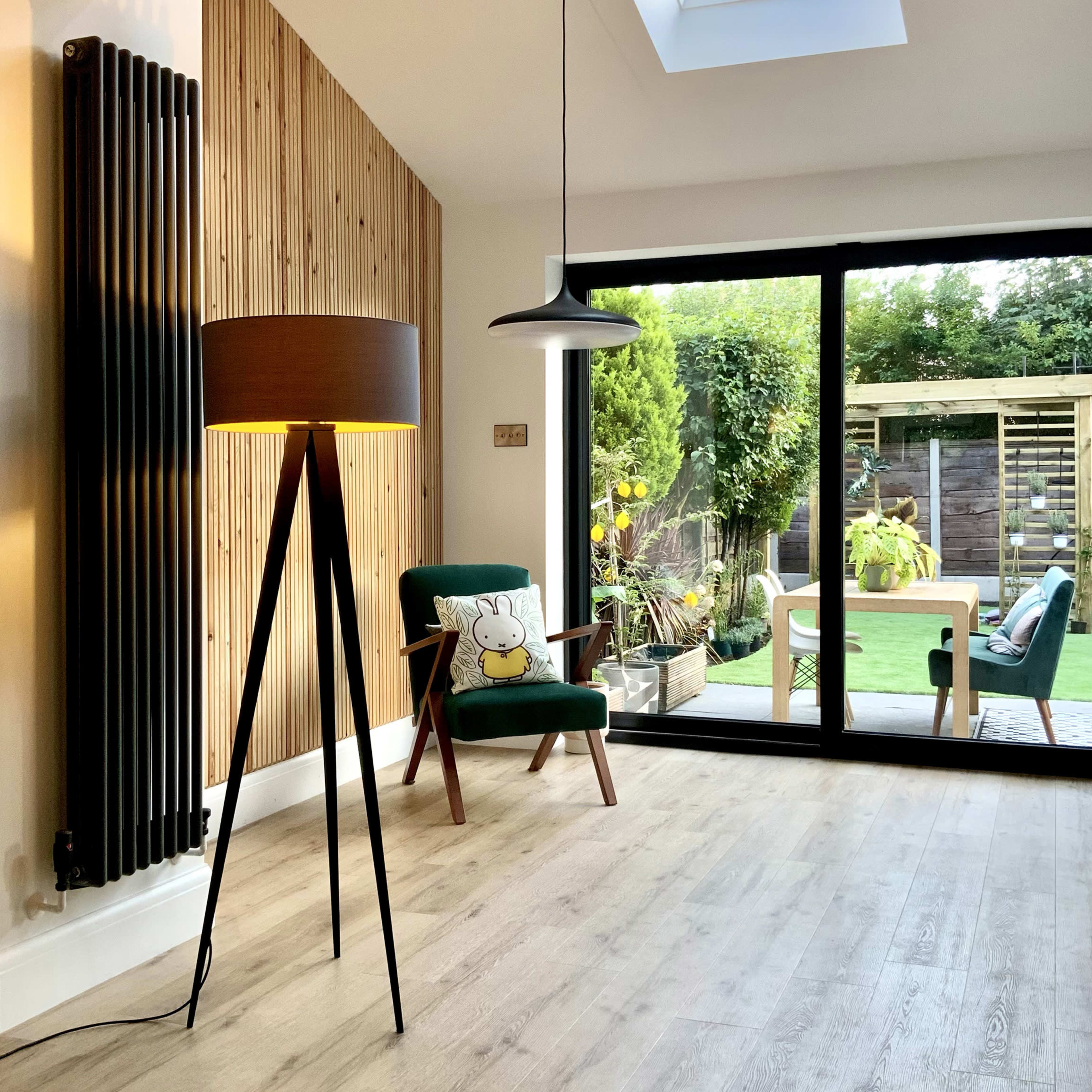 A modern living room features a tall, legged lamp and a green armchair with a decorative pillow, overlooking a garden through large glass doors.