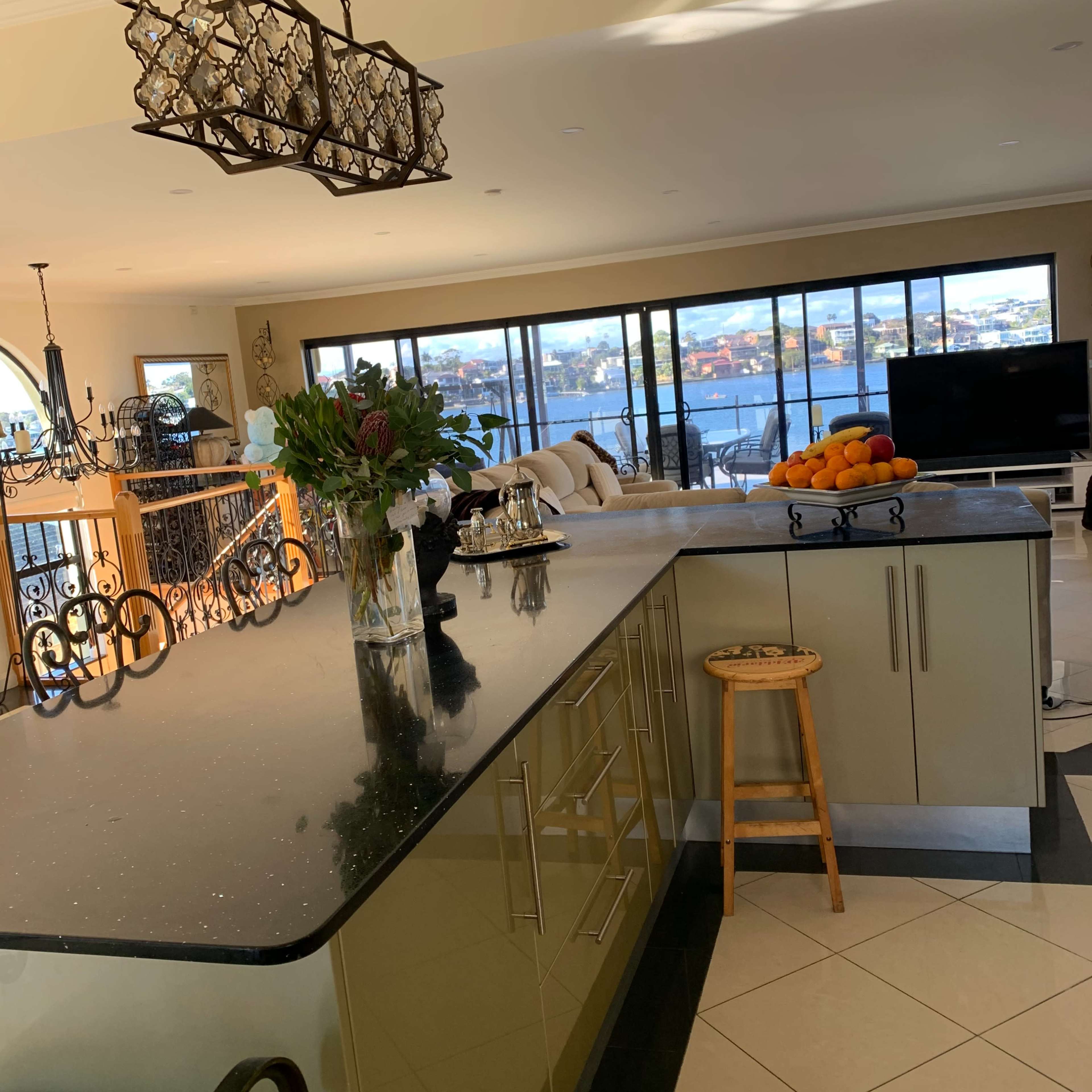 A modern kitchen with a dark countertop, a wooden stool, and large windows overlooking a waterfront view.