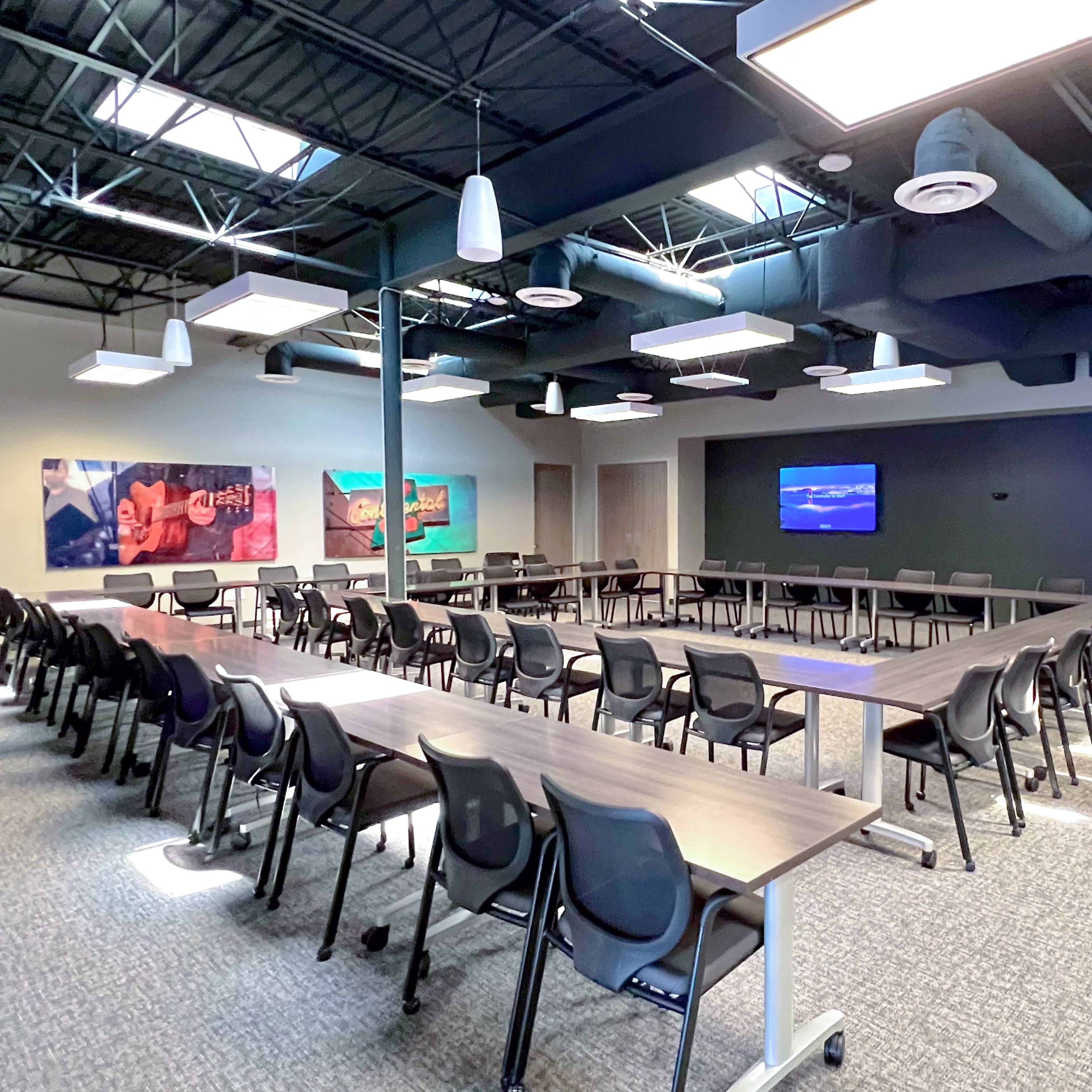 A spacious meeting room features multiple tables arranged in a U-shape, surrounded by black chairs, with two large screens displaying images on the walls.