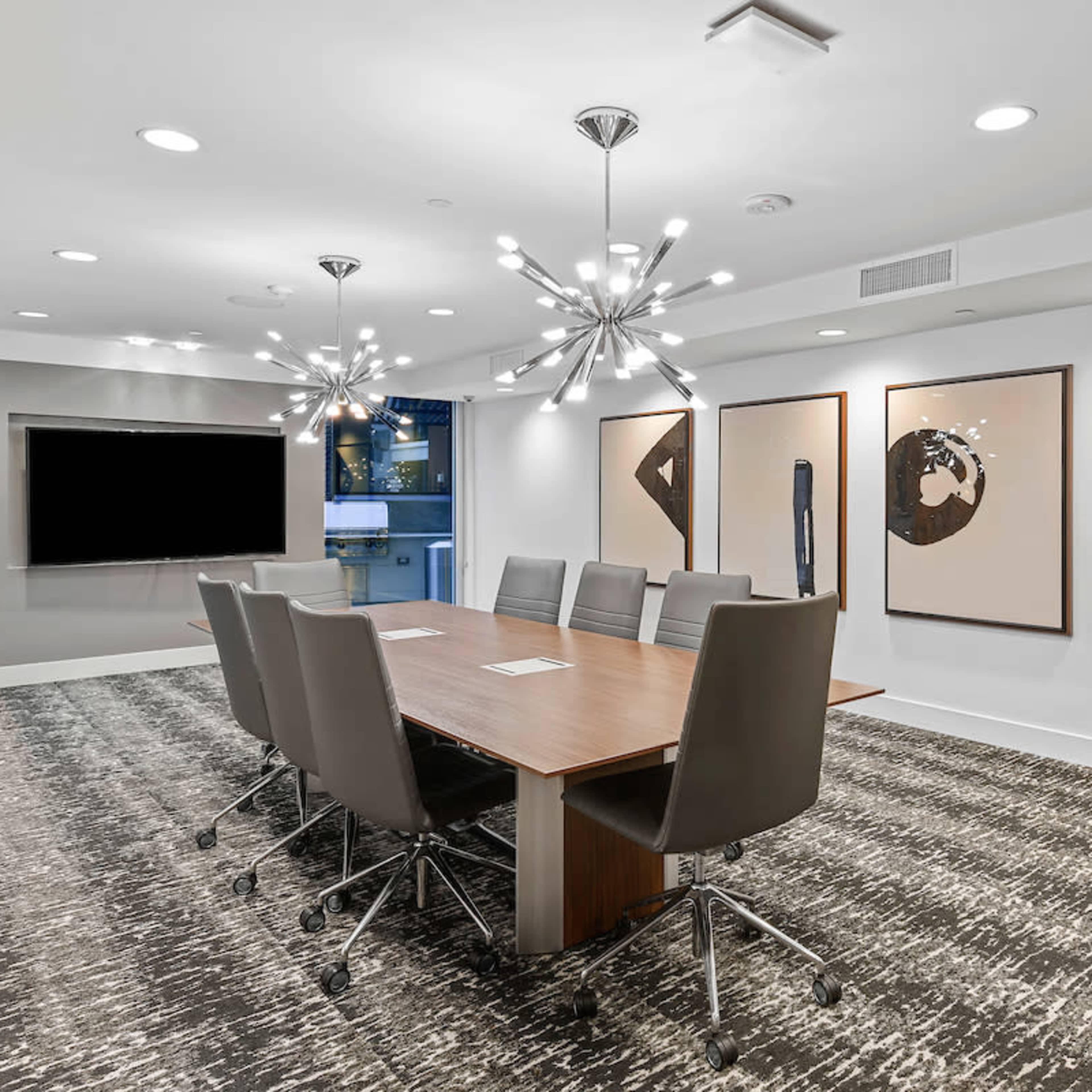 A modern conference room features a long wooden table surrounded by ergonomic chairs, with abstract artwork hanging on the walls and a large screen mounted for presentations.