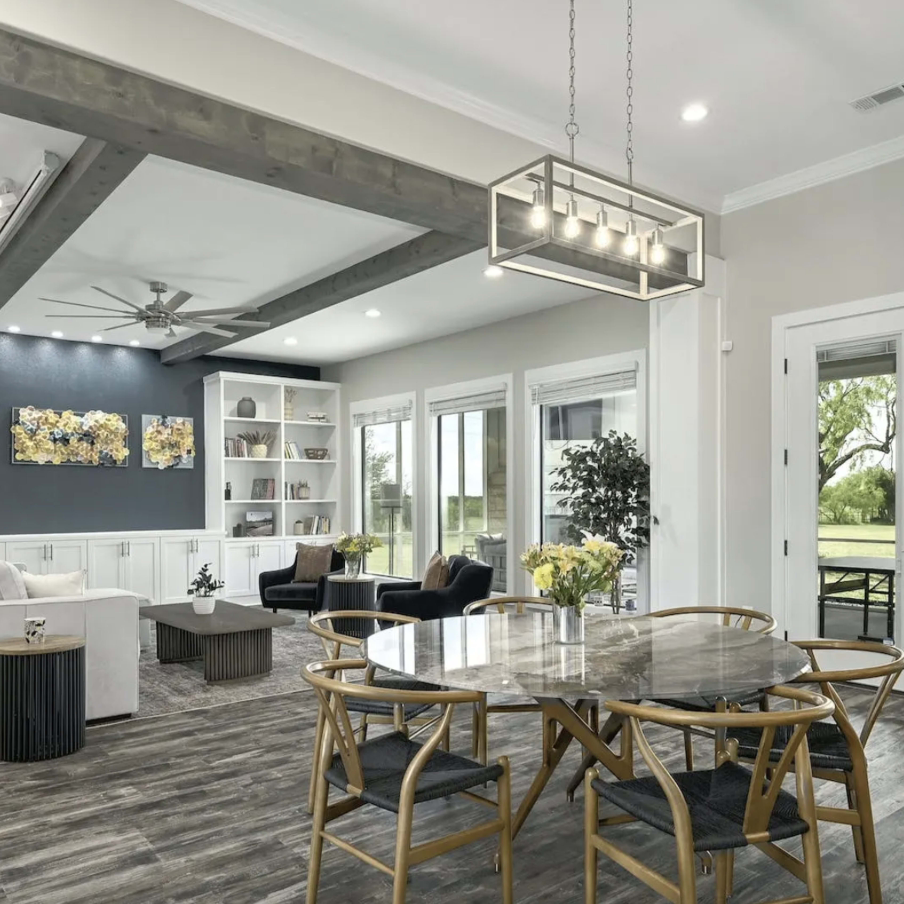 A modern living room and dining area with a light-colored sectional sofa, a circular dining table, and large windows that provide views of the outdoors.