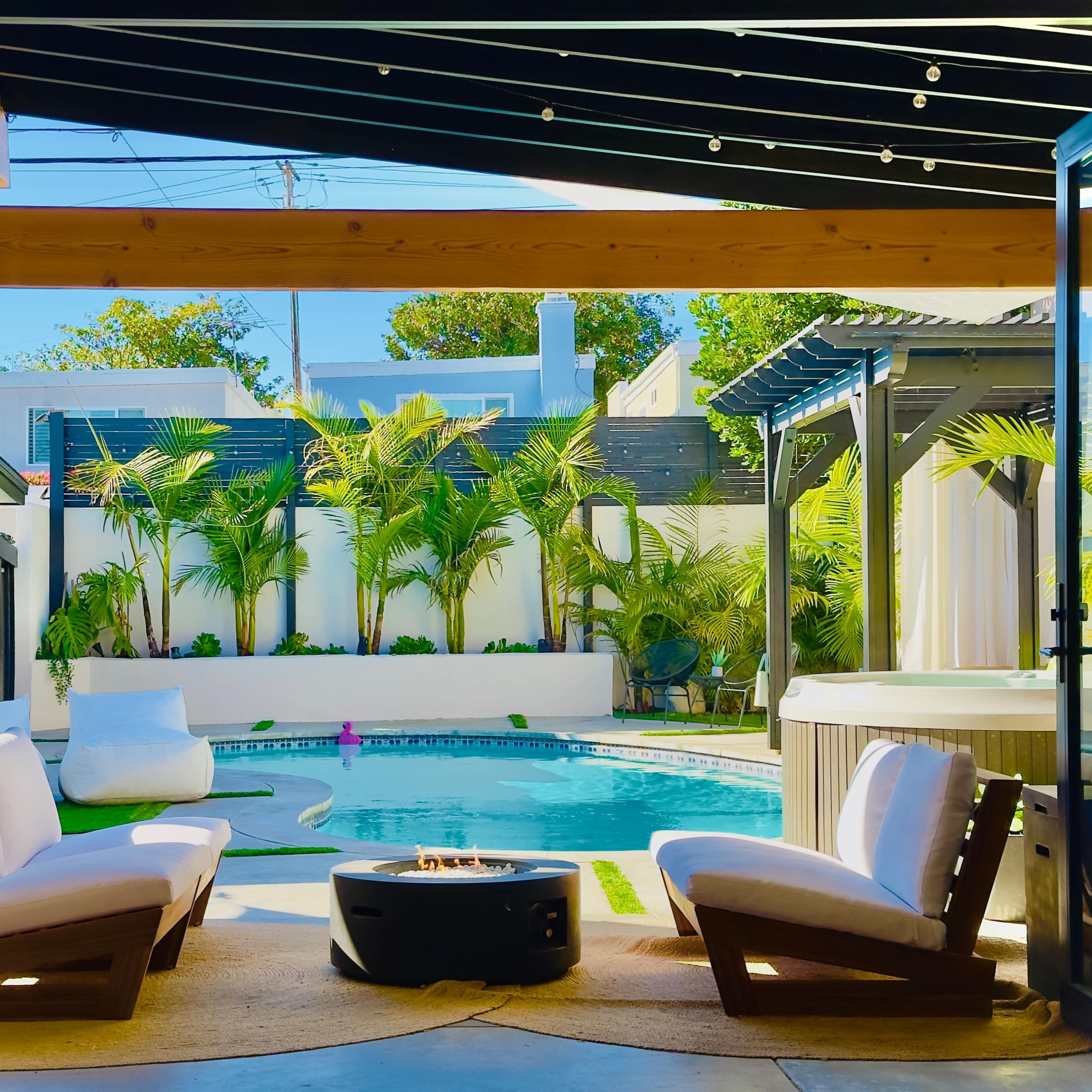 A modern outdoor seating area features two white chairs arranged around a fire pit, overlooking a pool surrounded by greenery.
