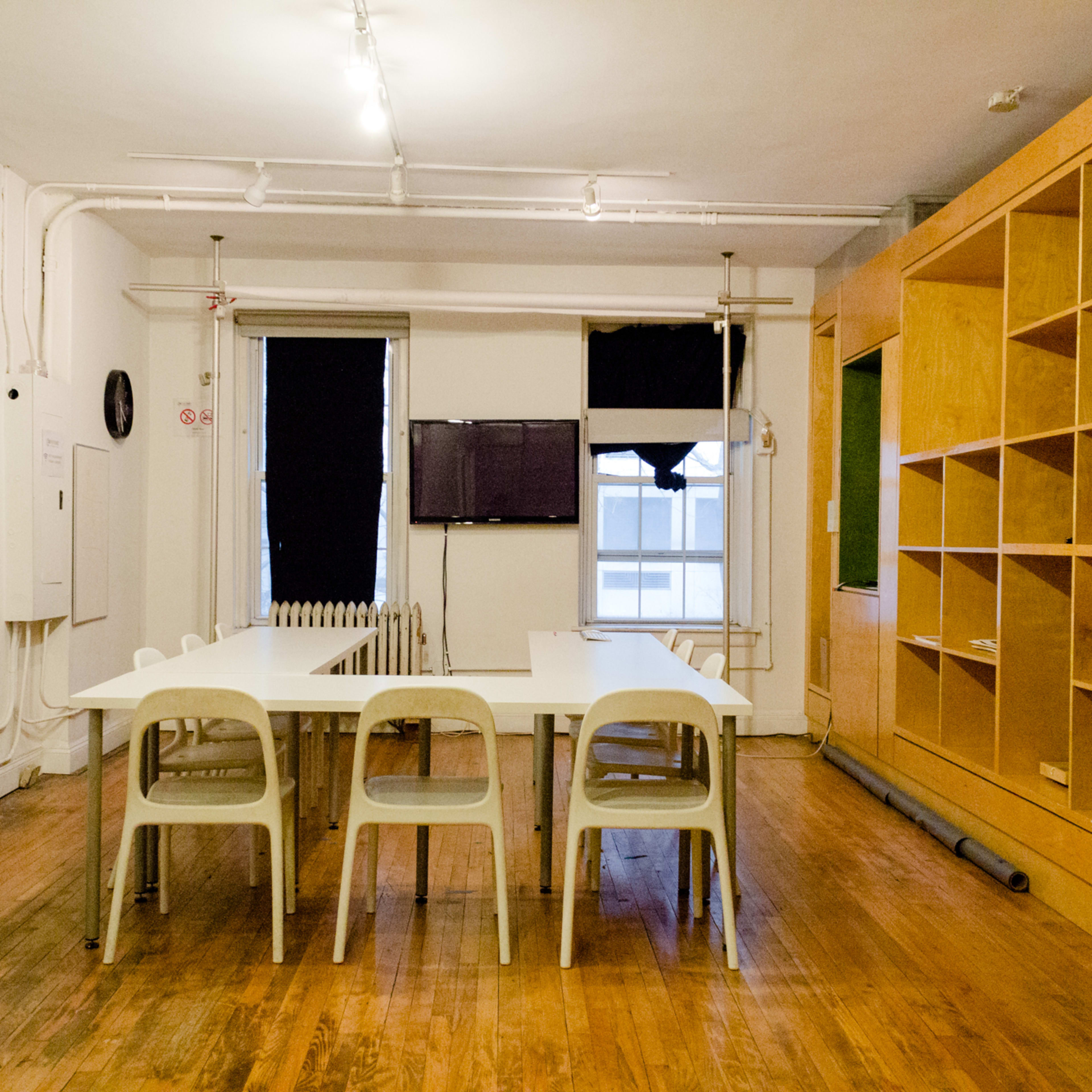 A spacious room features a long table surrounded by plastic chairs, with wooden shelves and a television mounted on the wall.