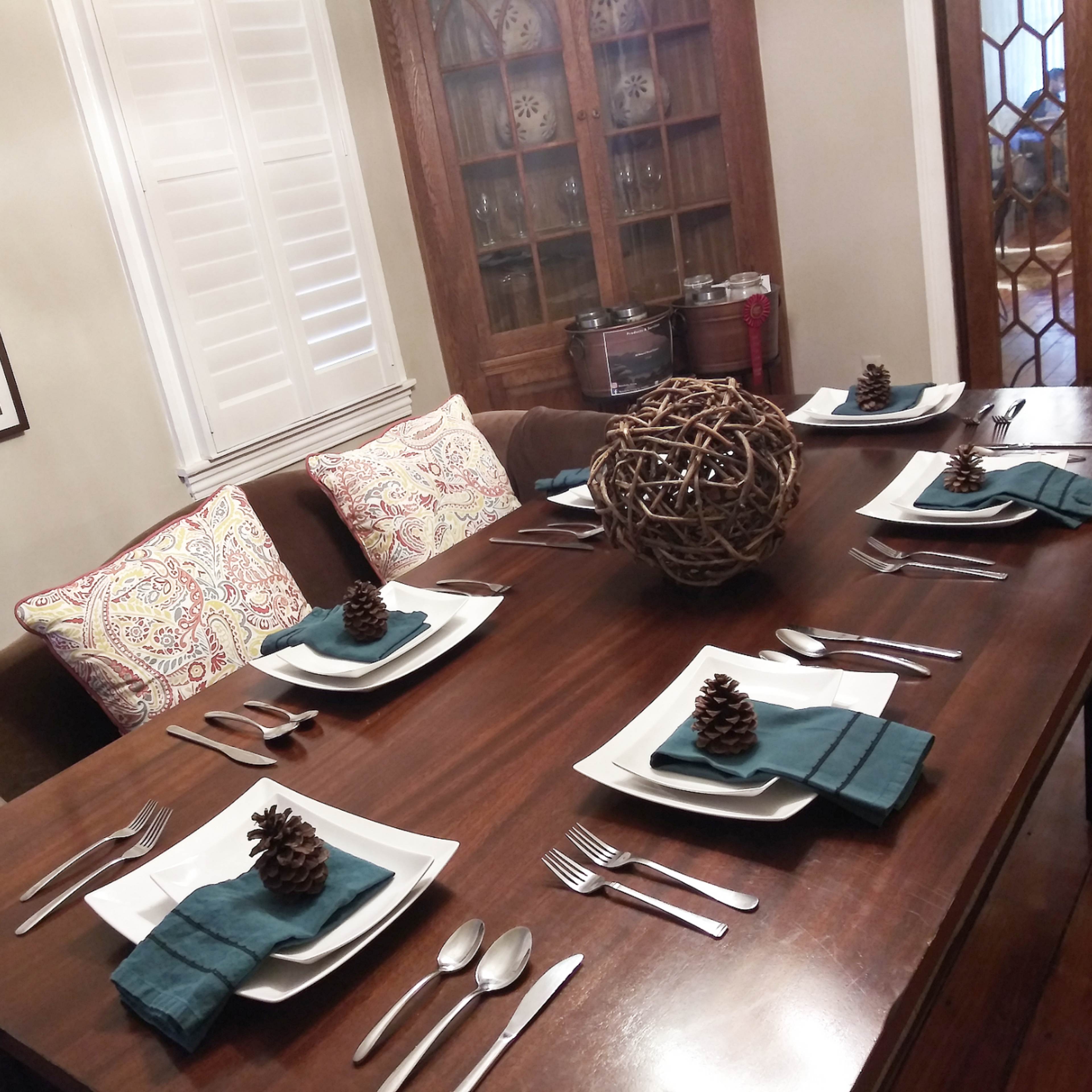 A dining table is set with plates, utensils, and decorative pinecones, surrounded by cushioned chairs and a cabinet with glassware in the background.