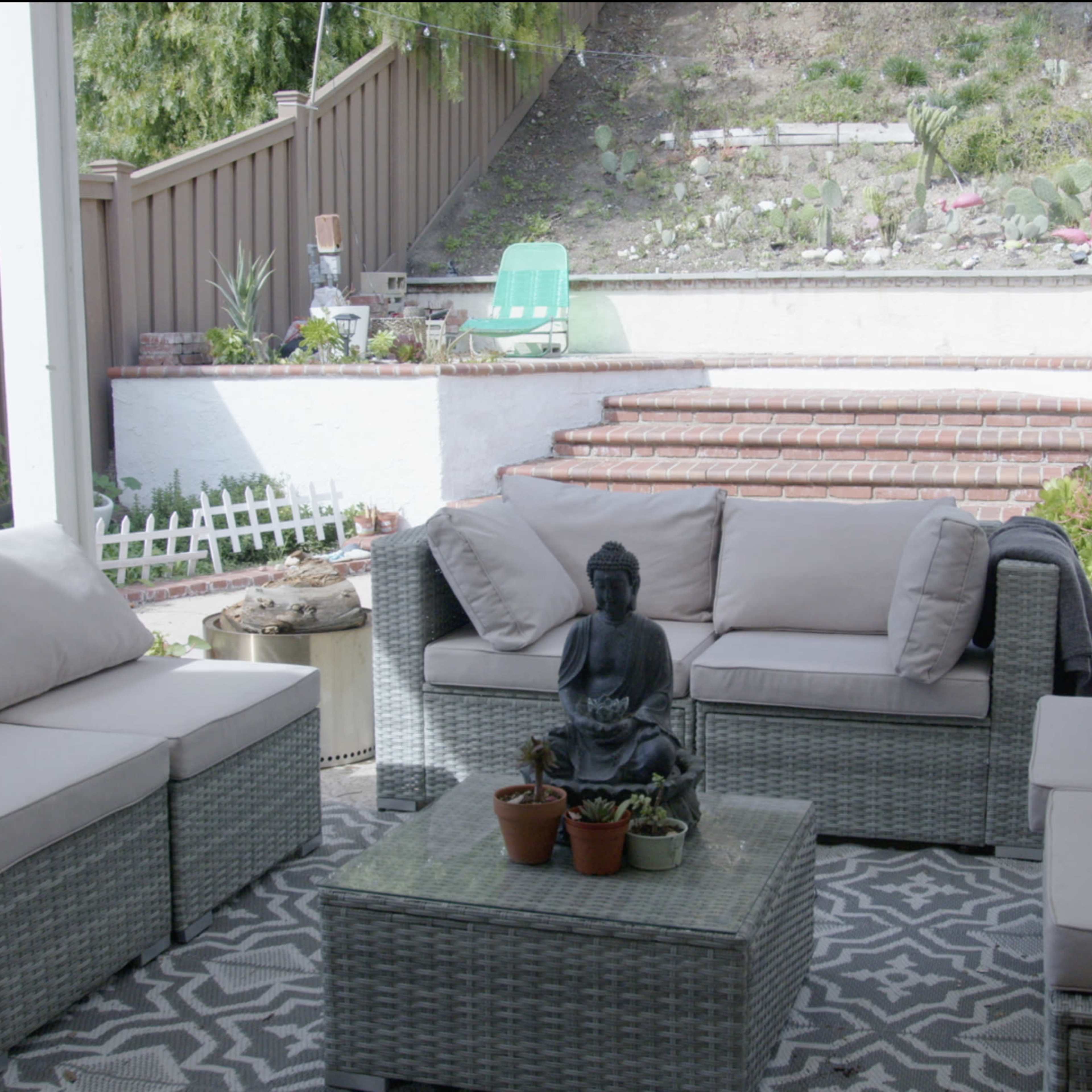 A patio area features a set of wicker furniture with light cushions arranged around a glass-top table, while a stone staircase leads to a landscaped yard in the background.