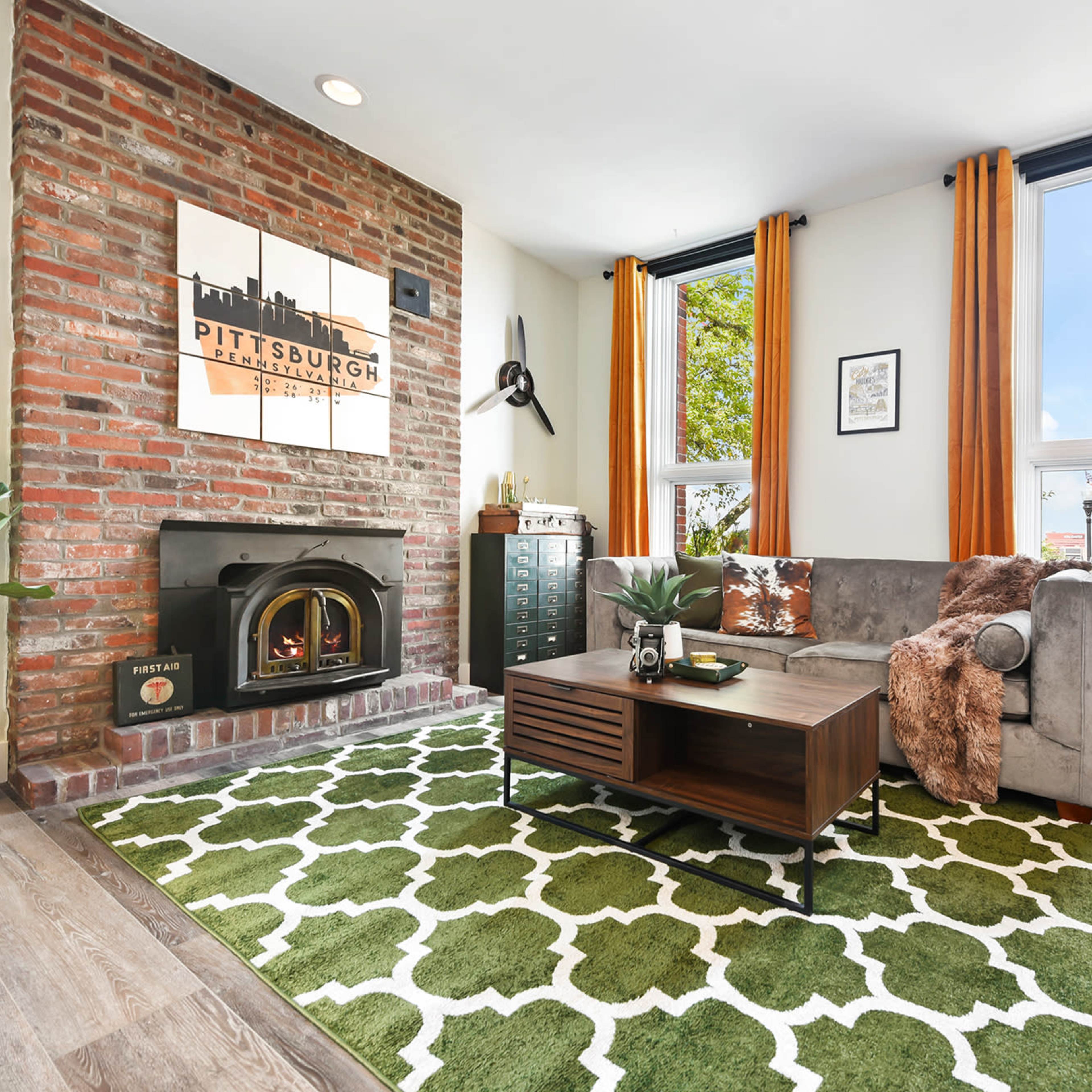 A modern living room with a brick wall featuring a Pittsburgh skyline artwork, a gray sofa, a wooden coffee table on a patterned rug, and large windows dressed with orange curtains.
