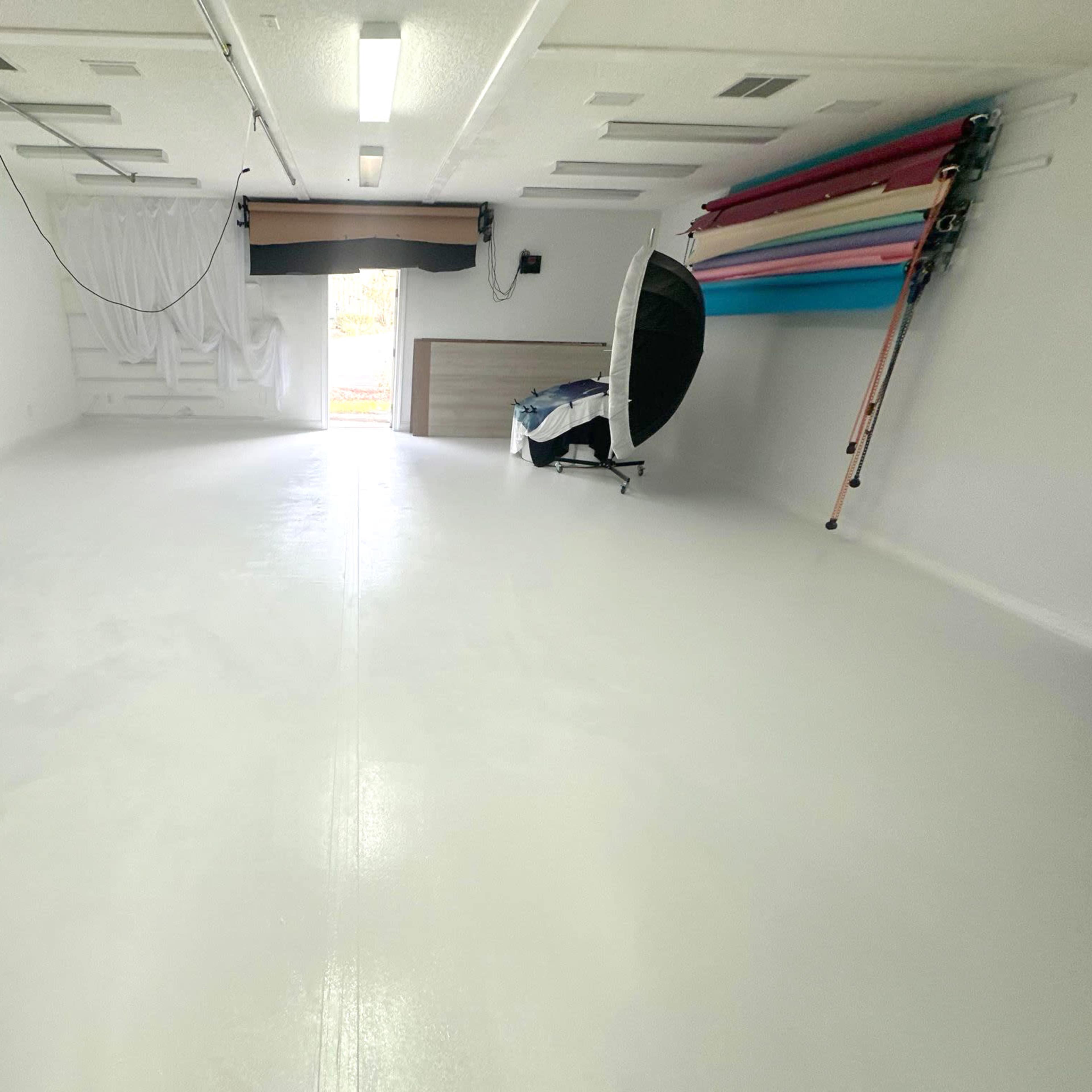 A large, empty, well-lit room with a smooth white floor and a backdrop stand against one wall.