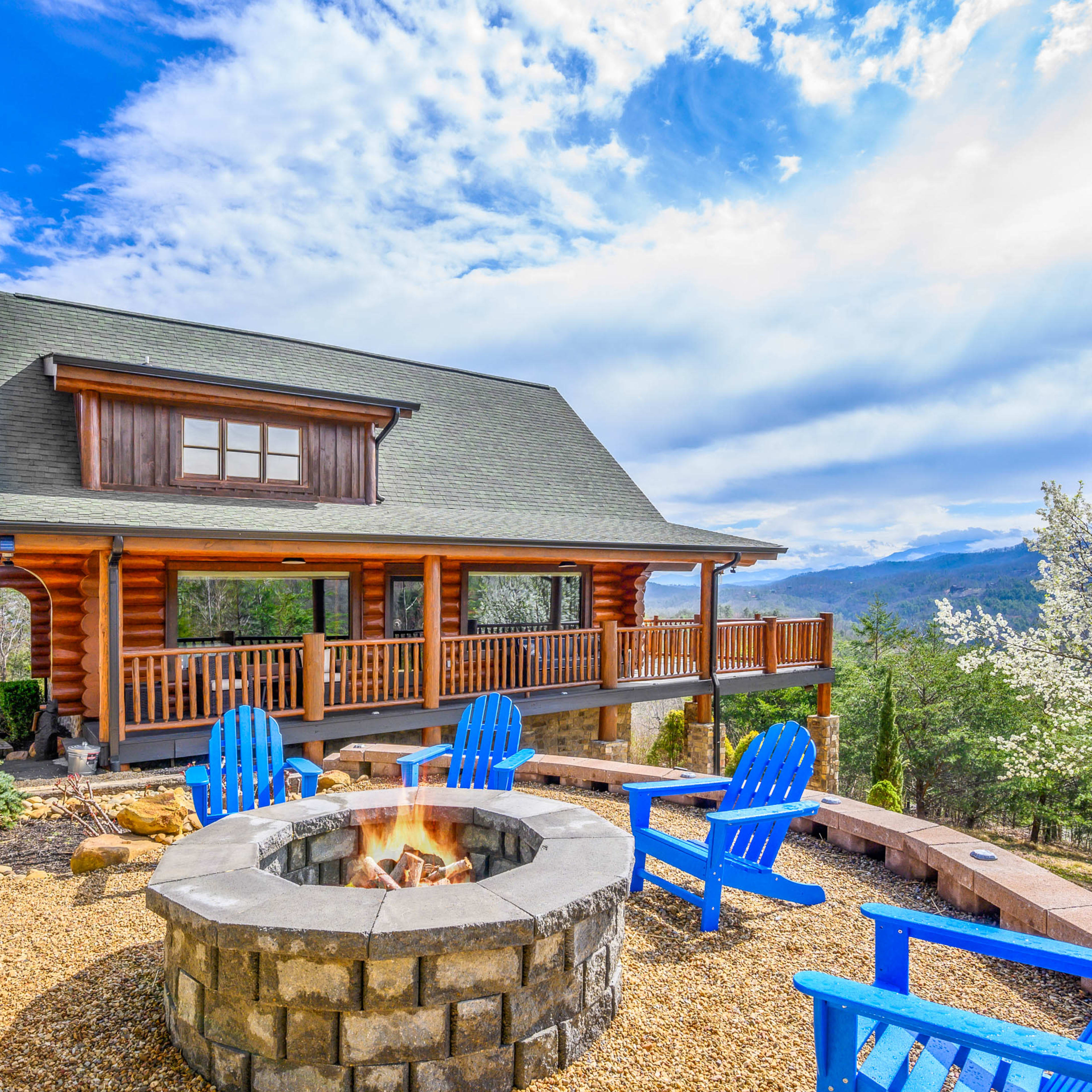 A log cabin with a spacious porch overlooks a fire pit surrounded by blue Adirondack chairs and a scenic landscape.
