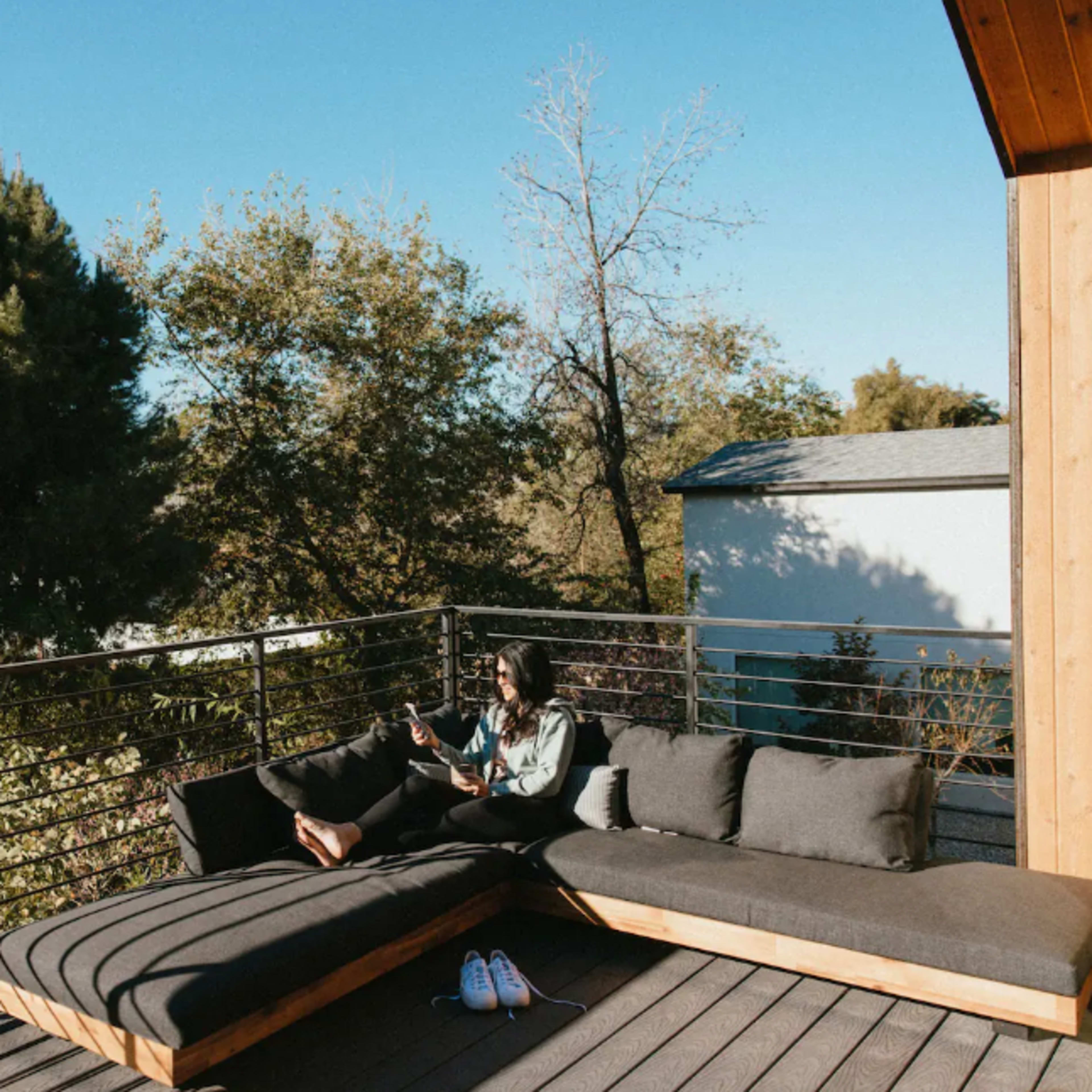 A person sits on a balcony couch, reading a book surrounded by trees and a clear blue sky.