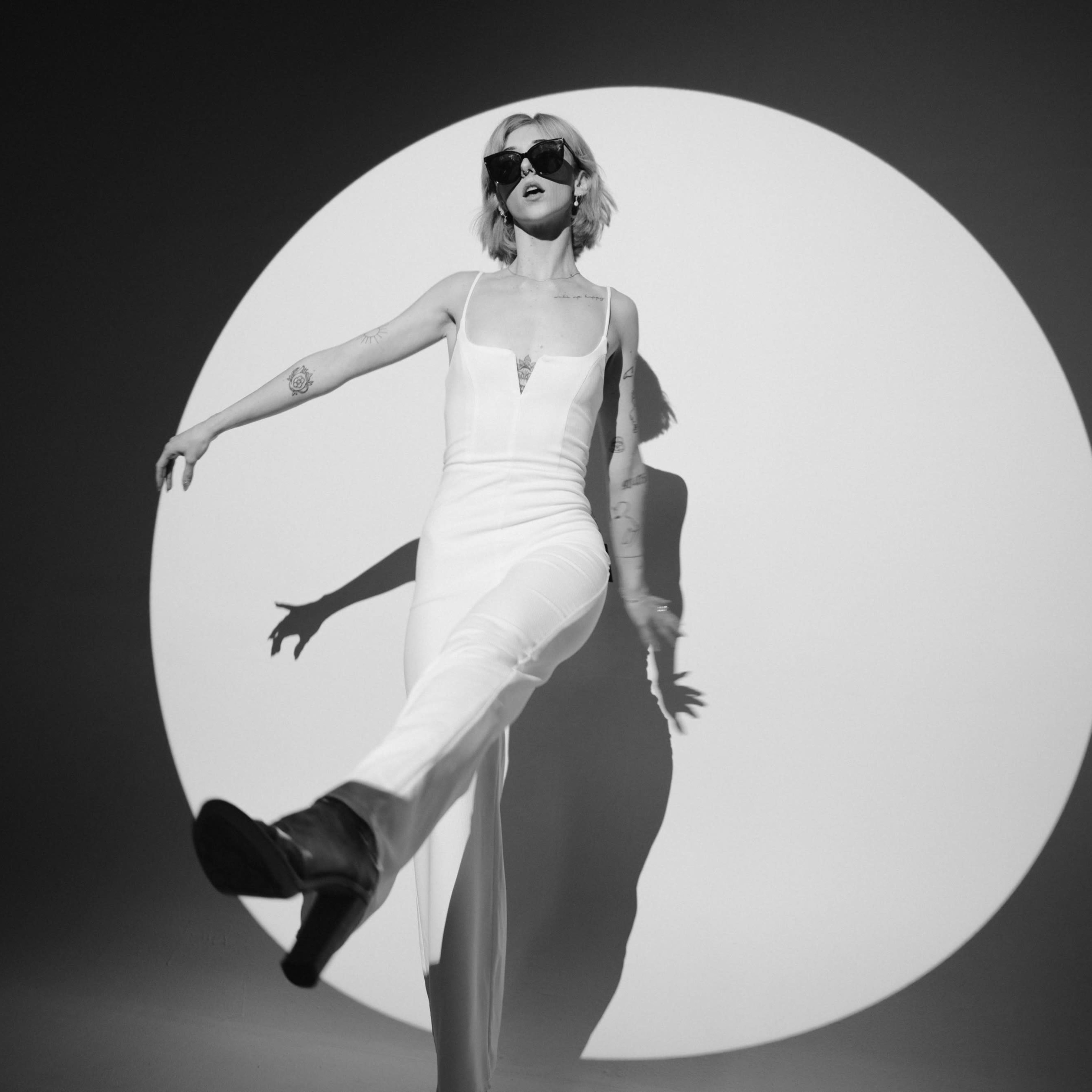 A person in a white outfit strikes a pose with one leg extended in front of a large circular backdrop.