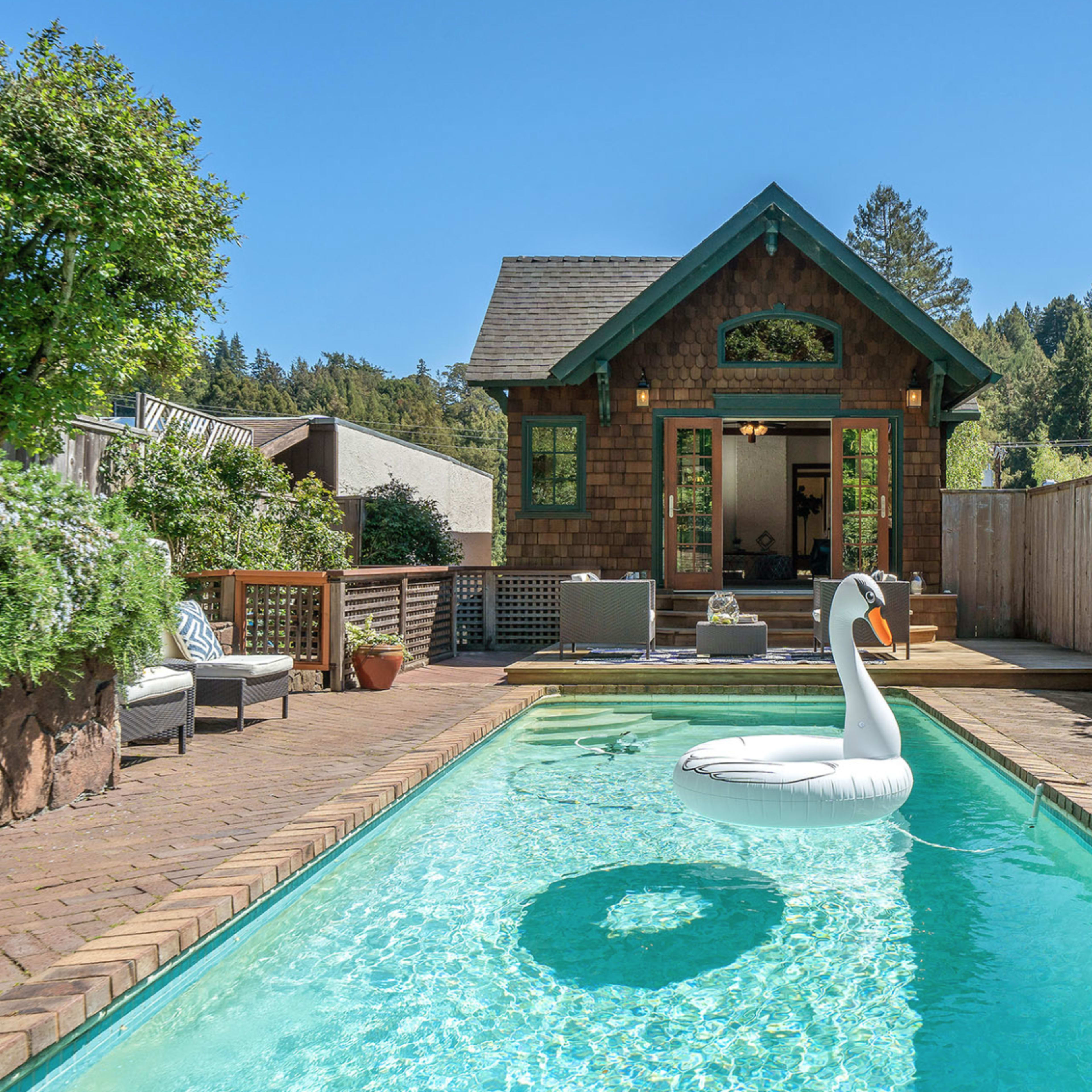 A rectangular swimming pool with a swan-shaped inflatable float sits in a backyard surrounded by greenery, leading to a house with a gabled roof and large windows.