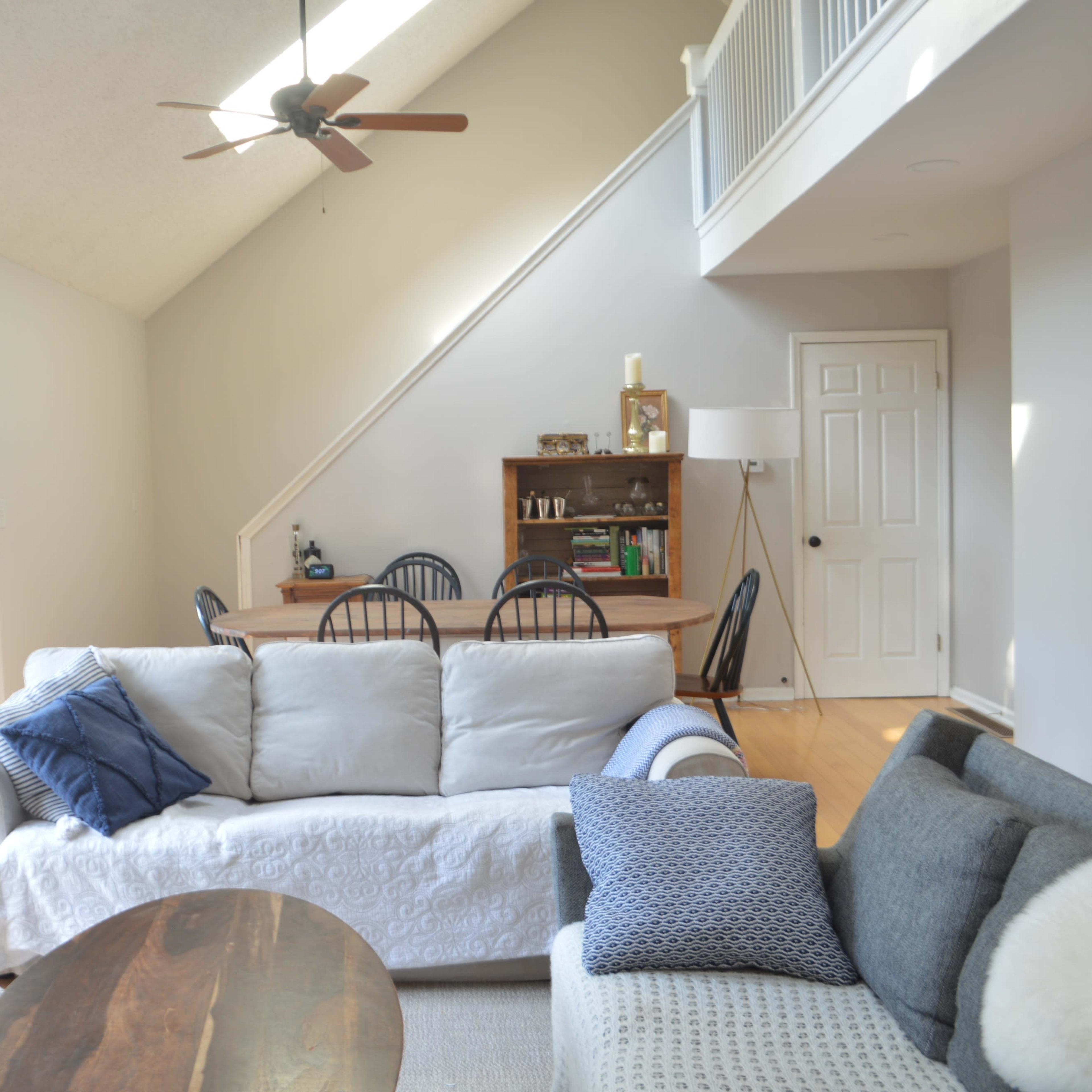 A bright living room features a light-colored sofa, a dark sofa, and a dining area with a wooden table and chairs, all illuminated by natural light from large sliding glass doors.