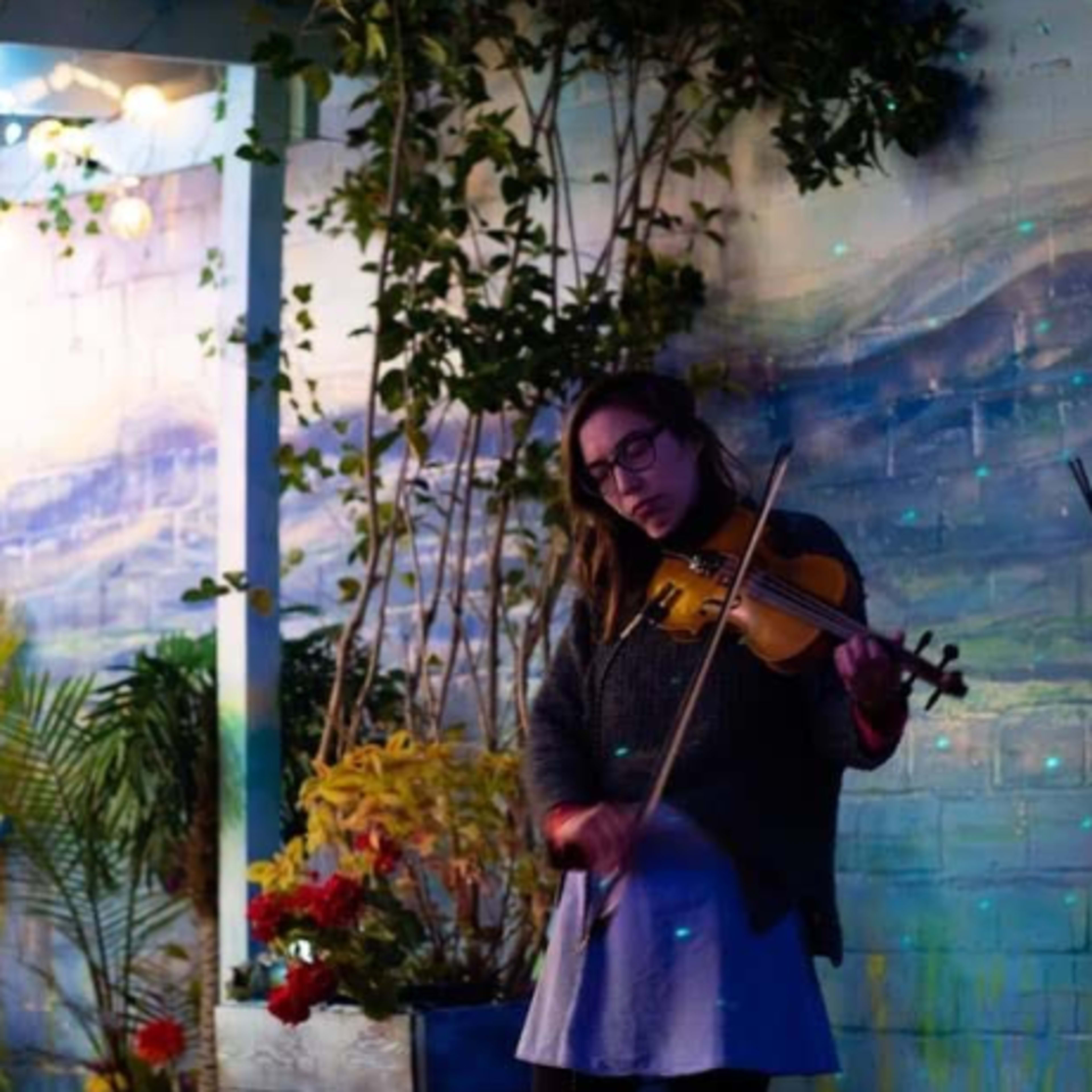 A woman plays a violin in front of a painted mural surrounded by plants and colorful lighting.