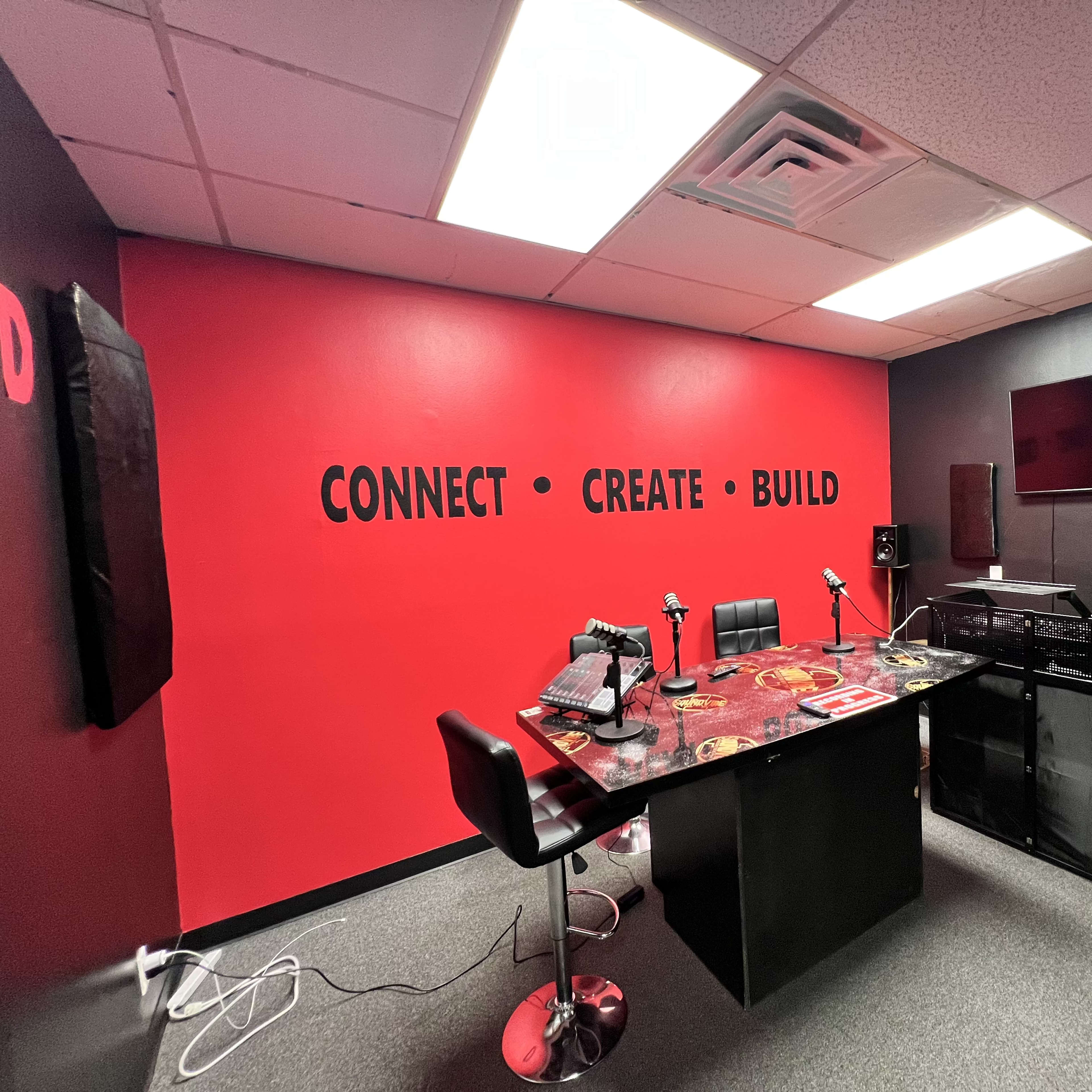 A recording studio with a red and black color scheme, featuring microphones on a table, chairs, and a television mounted on the wall.