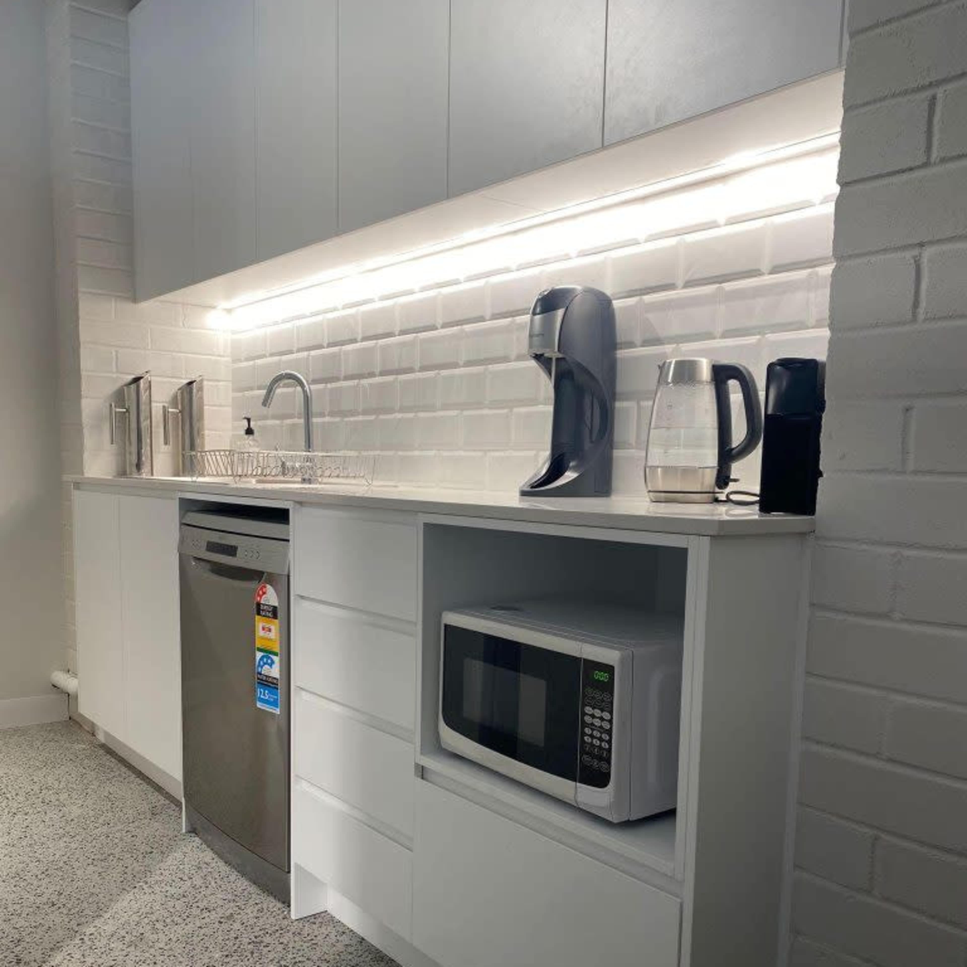 A modern kitchen countertop featuring a sink, a microwave in a cabinet, a coffee maker, and various kitchen appliances against a white tiled backsplash.