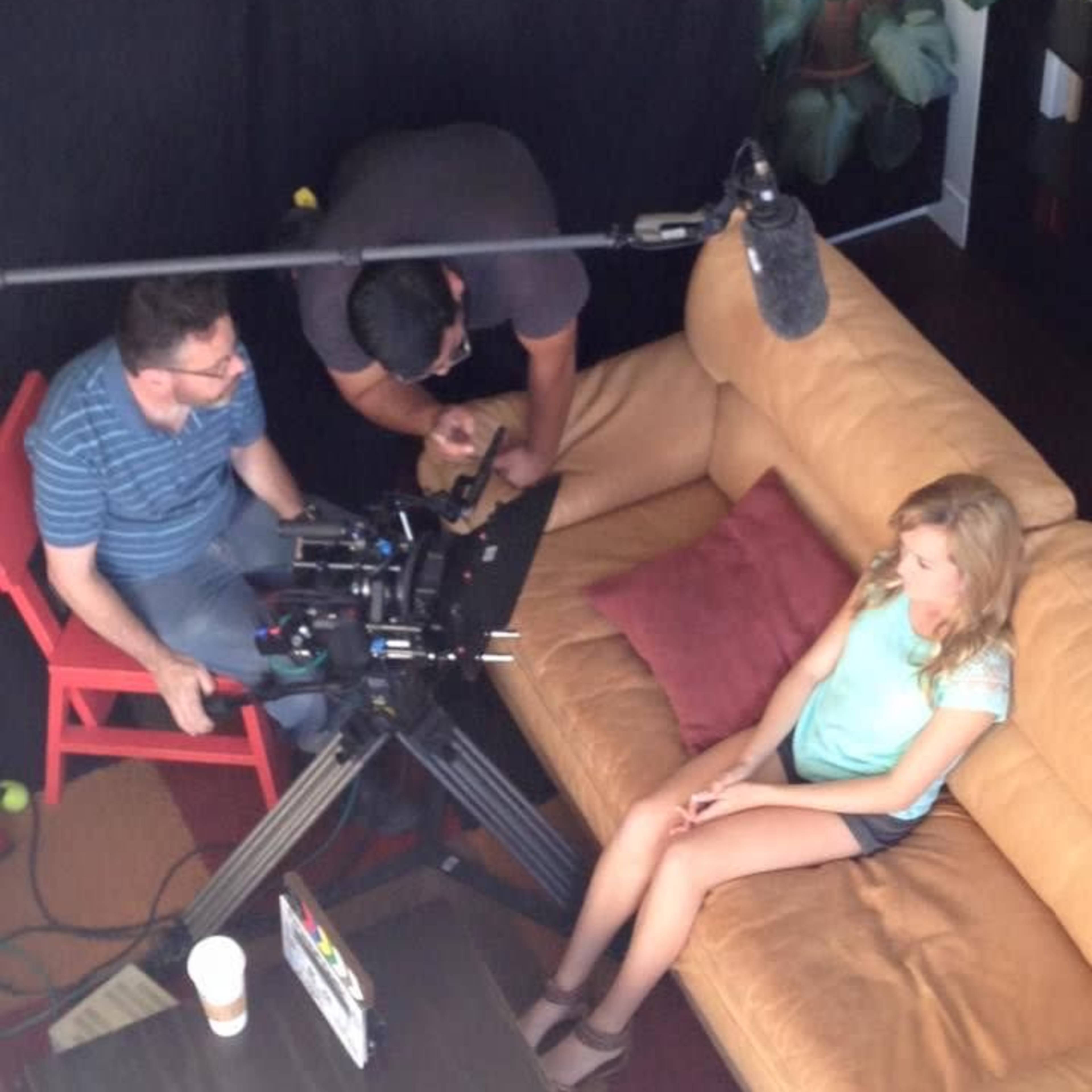 A film crew is setting up equipment around a seated woman in a living room with a couch and potted plants.