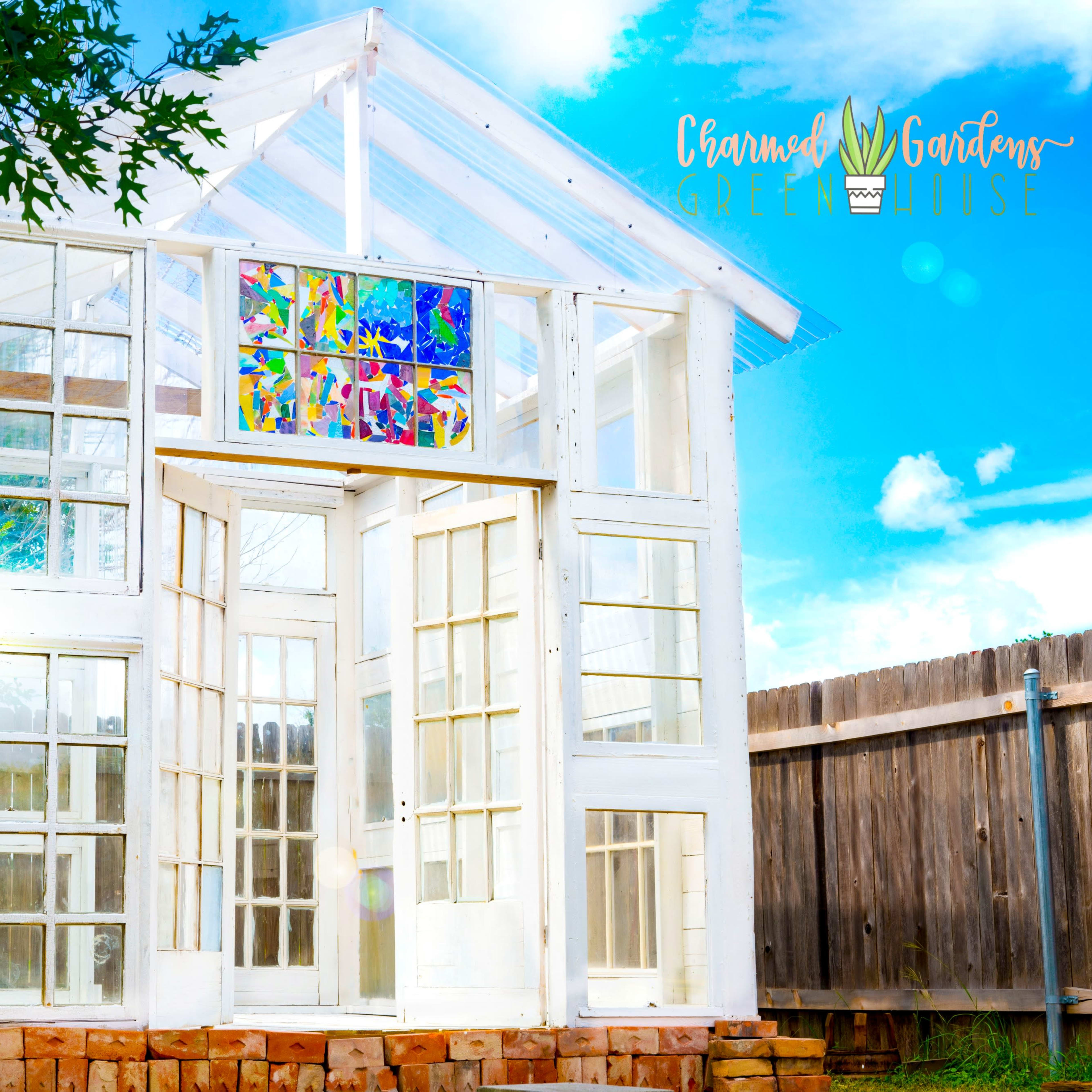 A white greenhouse constructed from glass panels, featuring a colorful stained glass window, situated in a yard surrounded by a wooden fence.