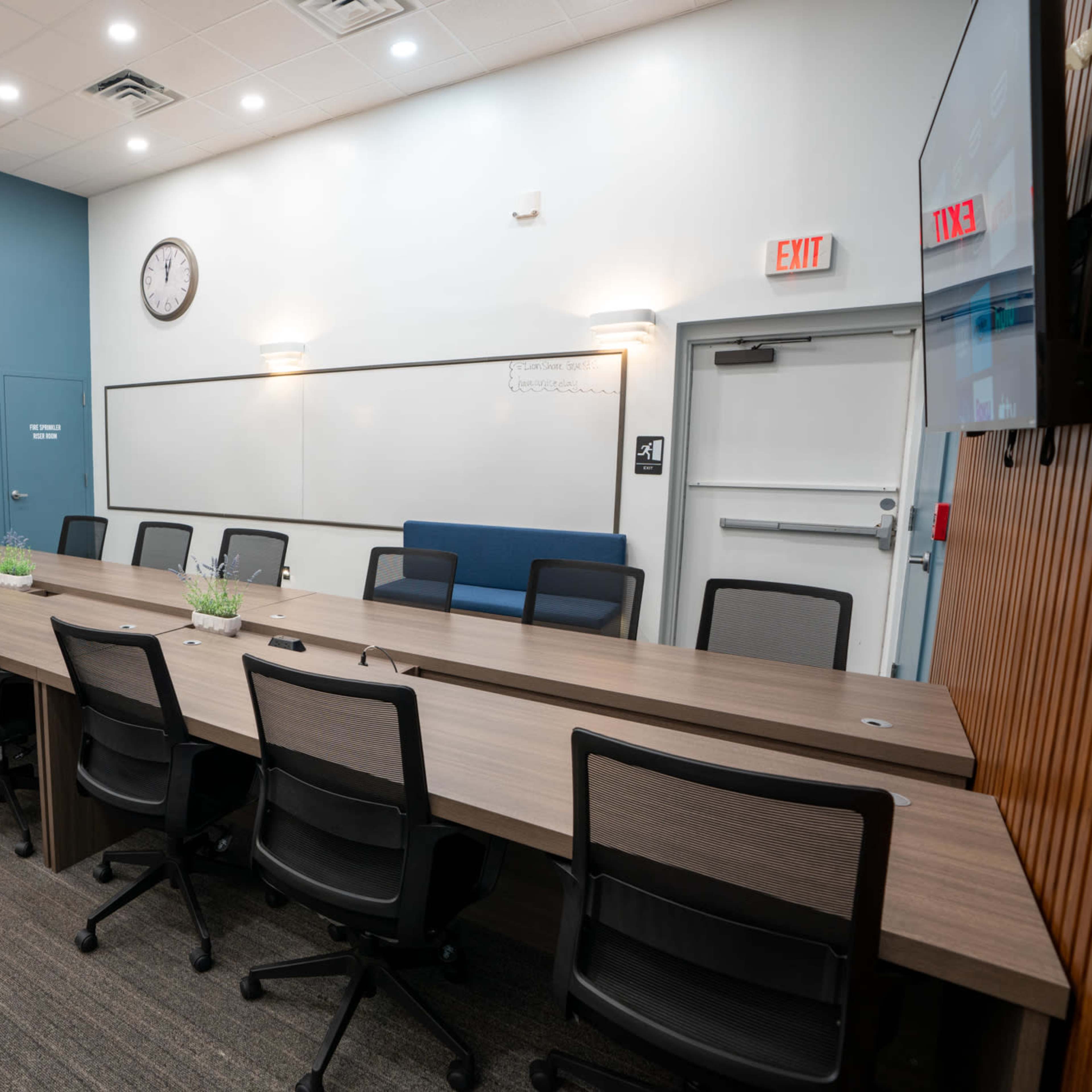 A conference room features a long, rectangular table with black chairs, a whiteboard, a clock on the wall, and a doorway leading to a restroom.