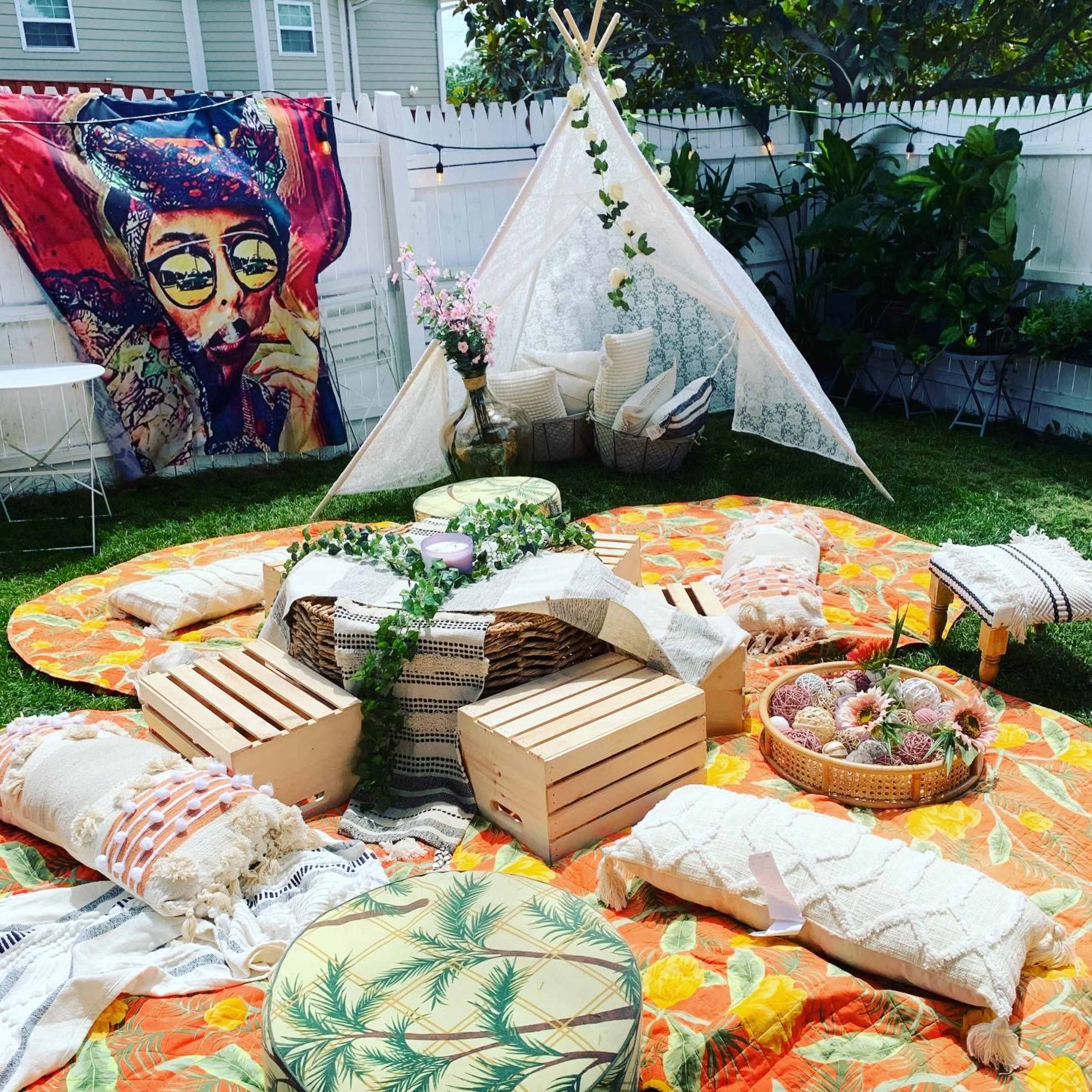A colorful outdoor setup with a teepee structure surrounded by cushions, crates, and decorative fabrics on a grassy area, with a vibrant tapestry hanging on the fence.