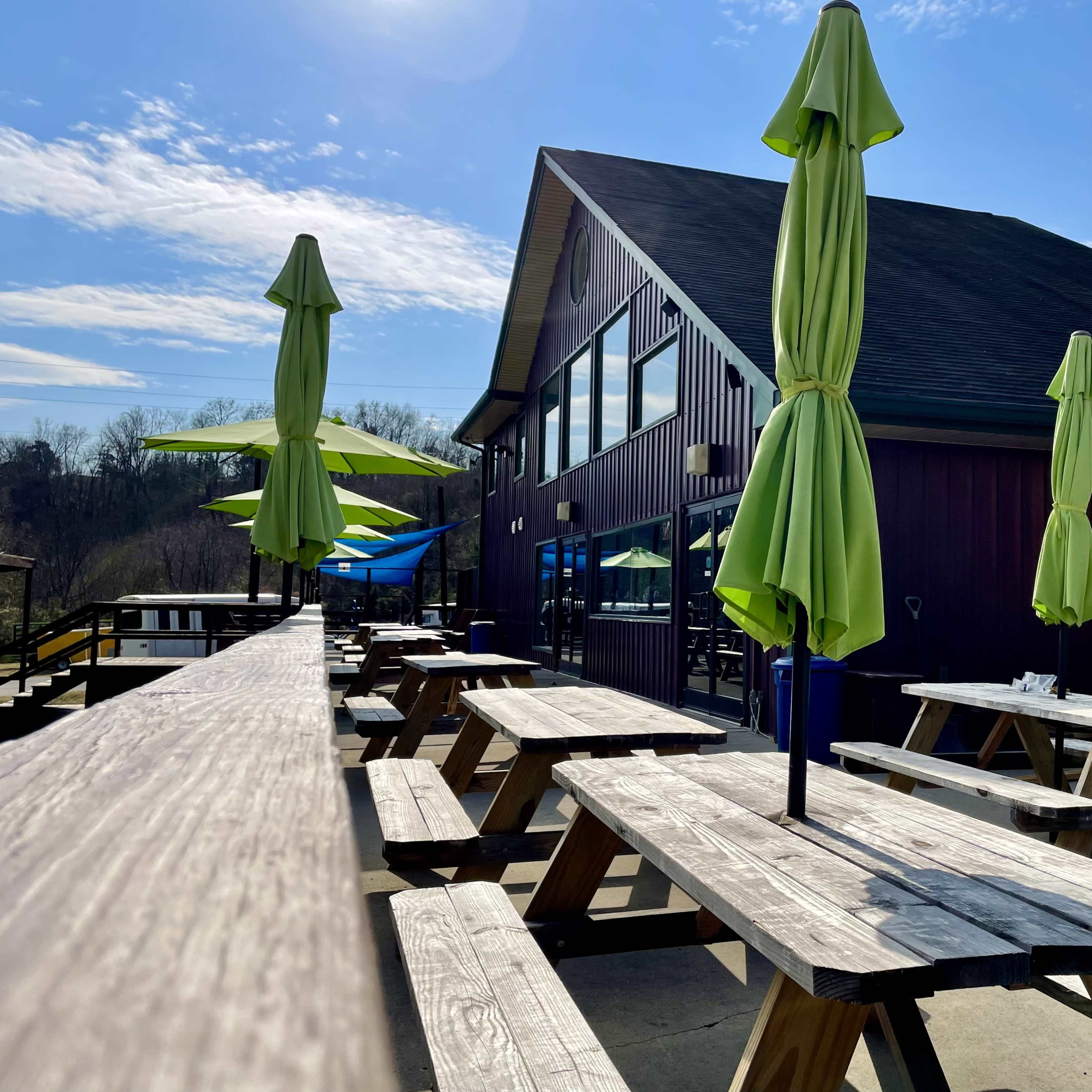 A series of wooden picnic tables with green umbrellas are arranged in front of a large building under a clear blue sky.
