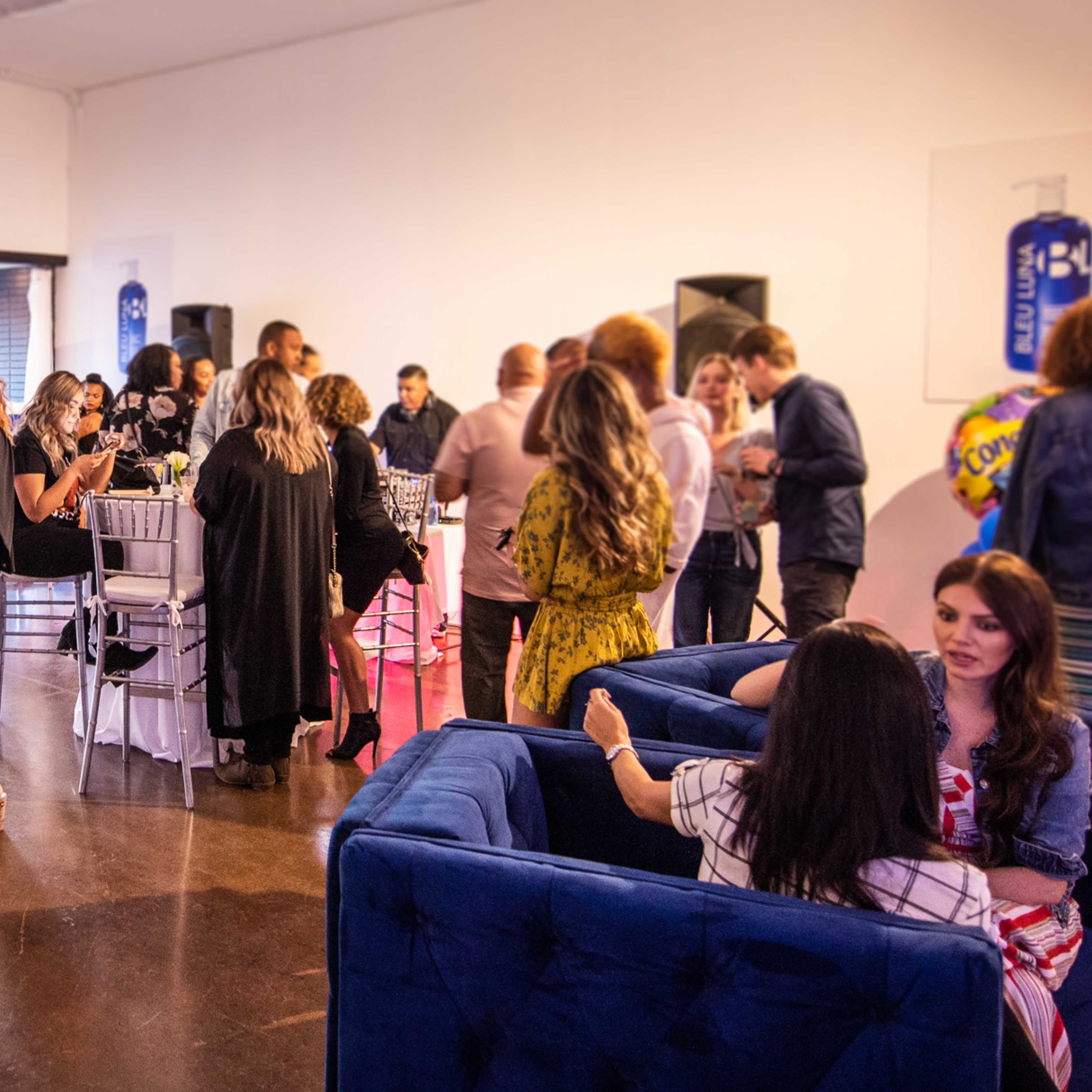 A lively gathering of people mingles at a spacious indoor venue with tables and a blue seating area.