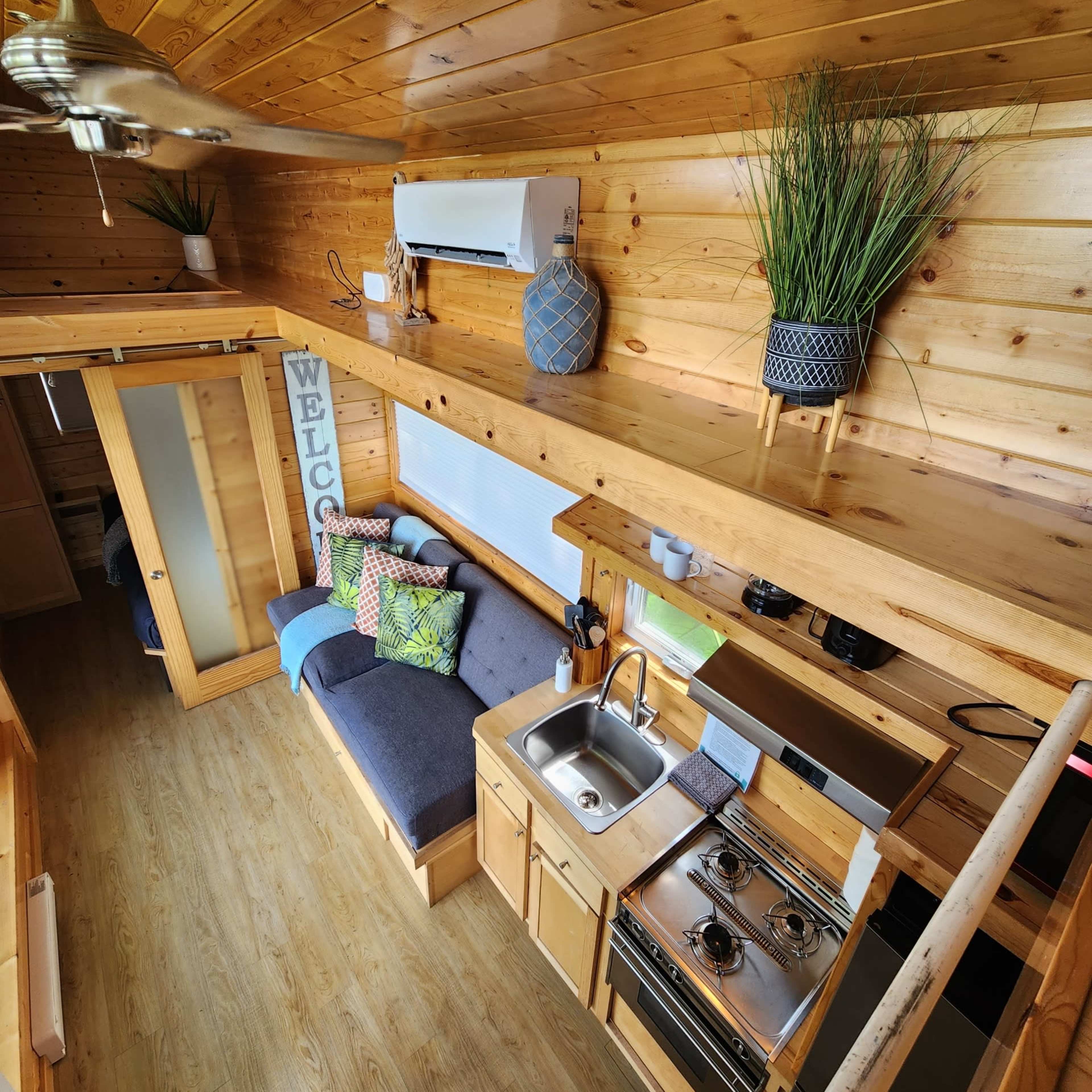 A compact wooden interior of a tiny home featuring a kitchenette, seating area, and decorative plants on shelves.