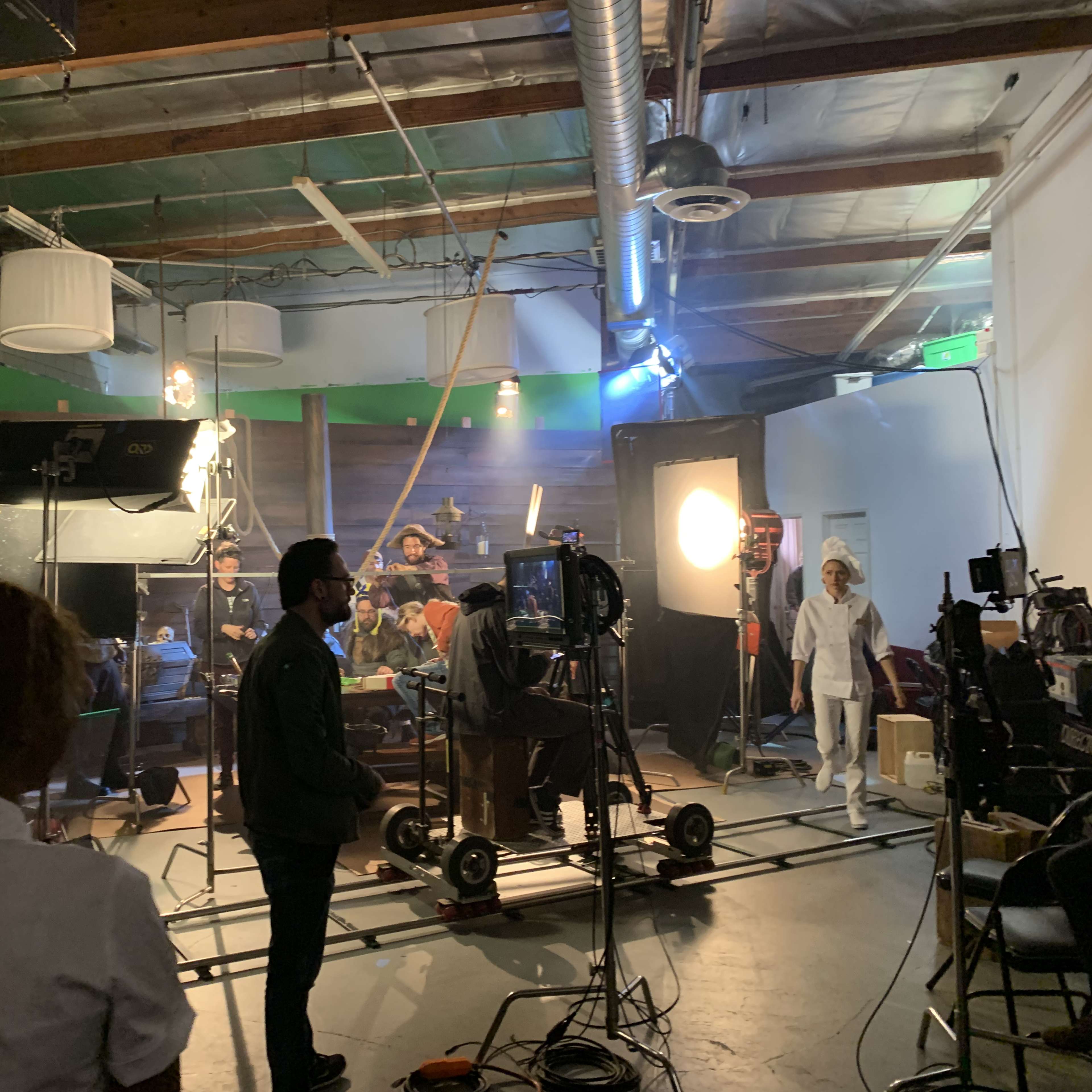 A film crew is set up in a studio with green screen backgrounds and multiple cameras, lights, and chairs scattered around.