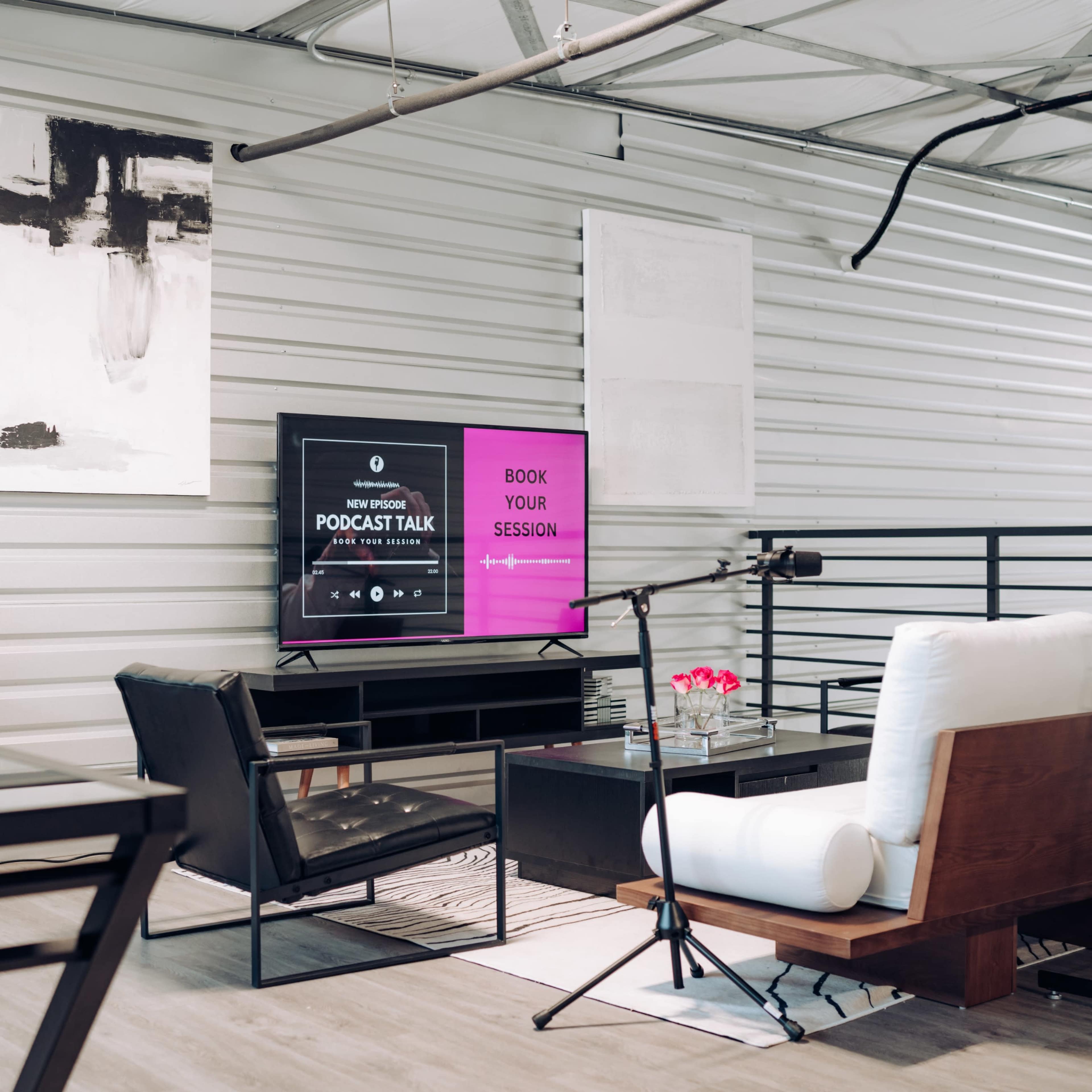 A modern podcasting space features a television displaying a booking interface, a comfortable seating area, and microphones set up for recording.