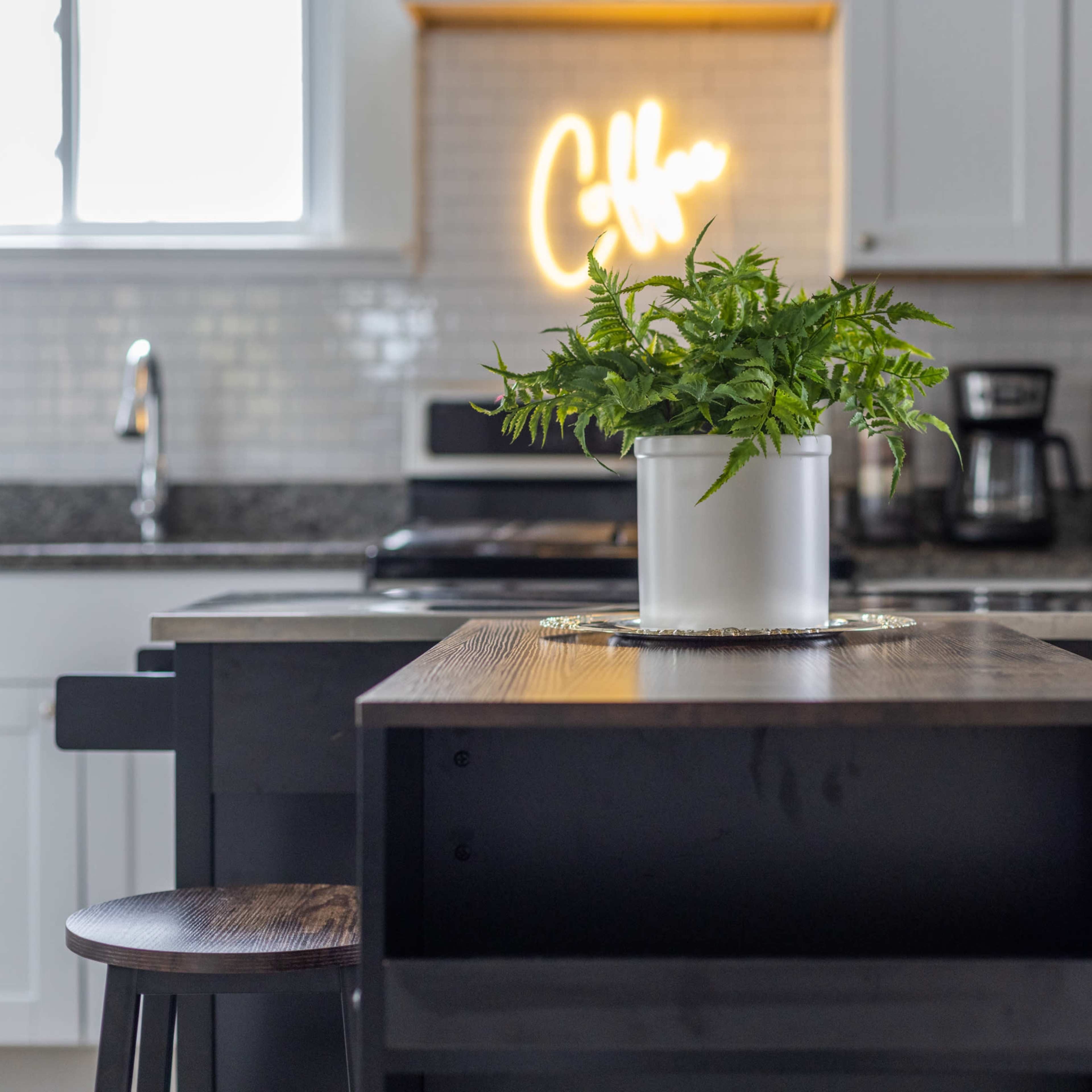 A kitchen scene features a wooden island with two stools, a potted plant in the center, and a glowing "Coffee" sign on the wall.
