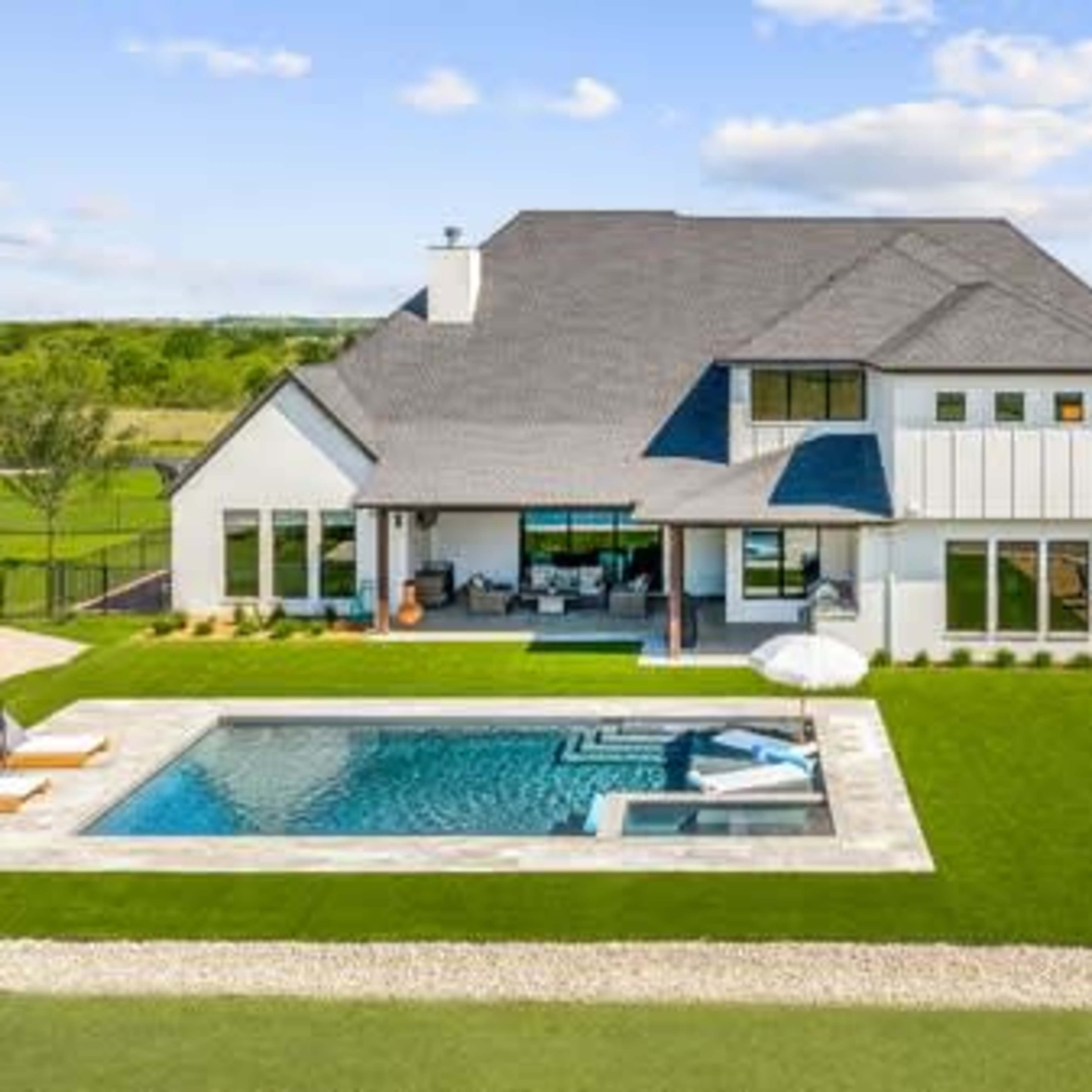 A modern two-story house with a swimming pool, outdoor seating area, and landscaped green lawn under a blue sky.