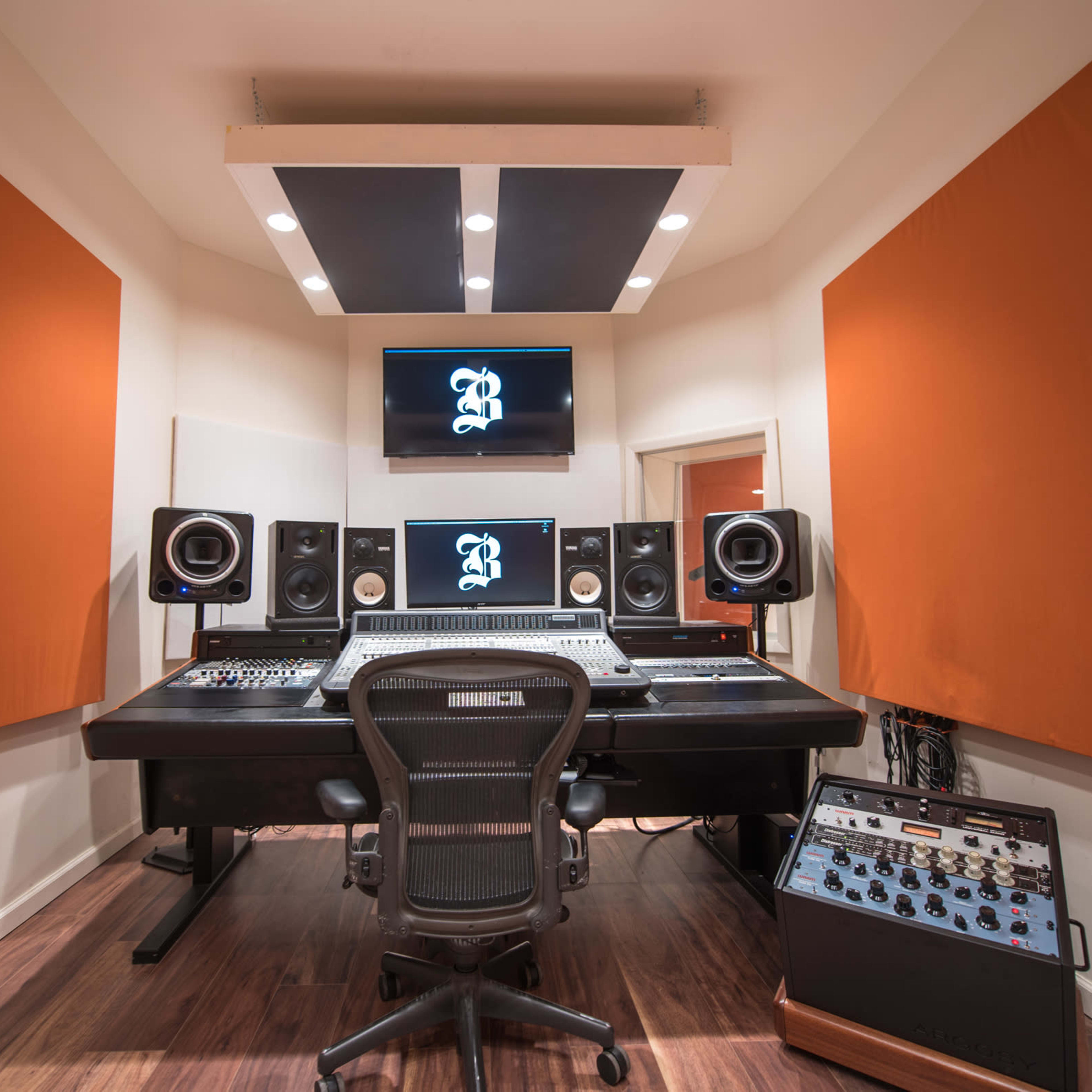 A recording studio with a control desk, large speakers, a TV monitor, and soundproofing panels on the walls.
