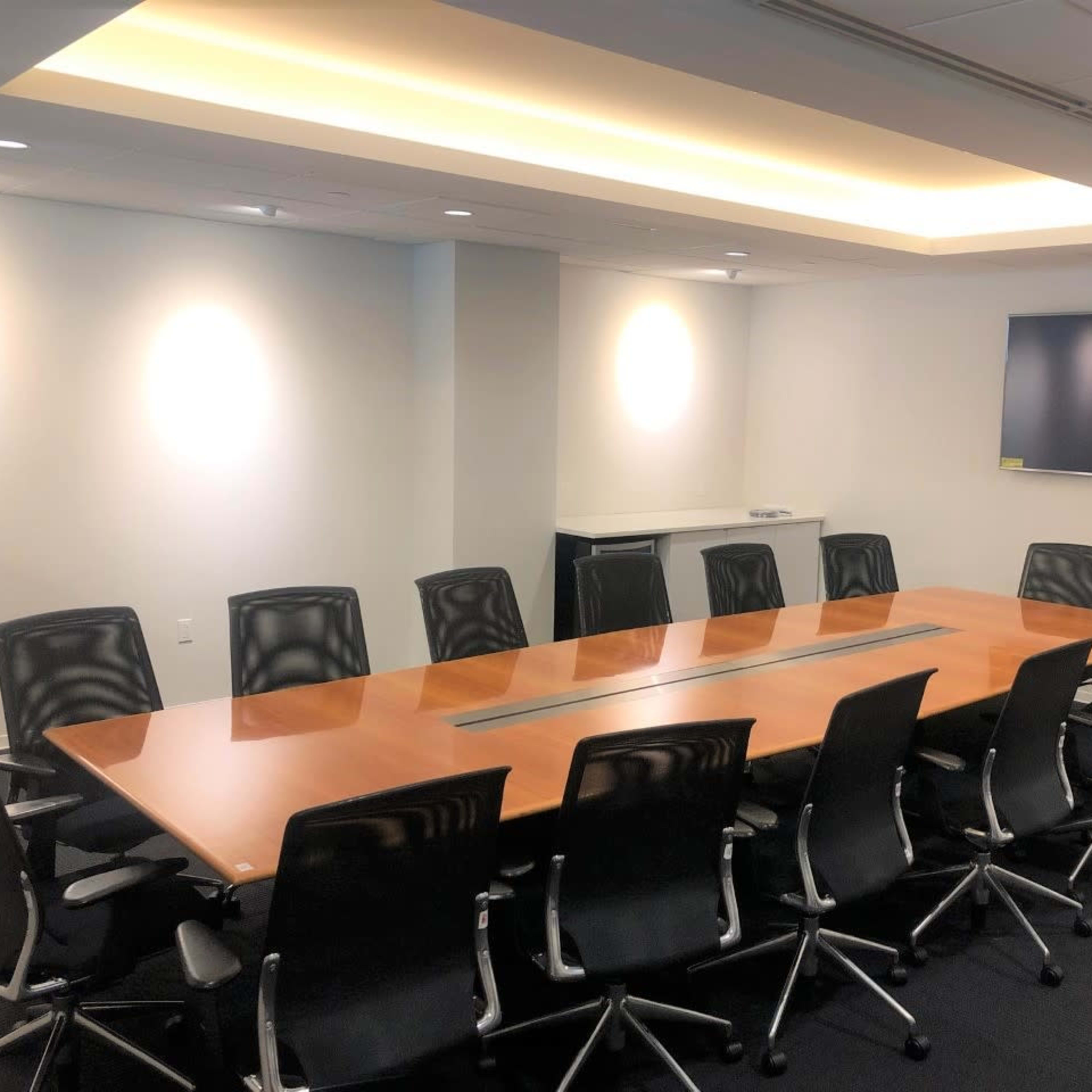 A large, rectangular conference table surrounded by black office chairs in a well-lit meeting room with a wall-mounted television.