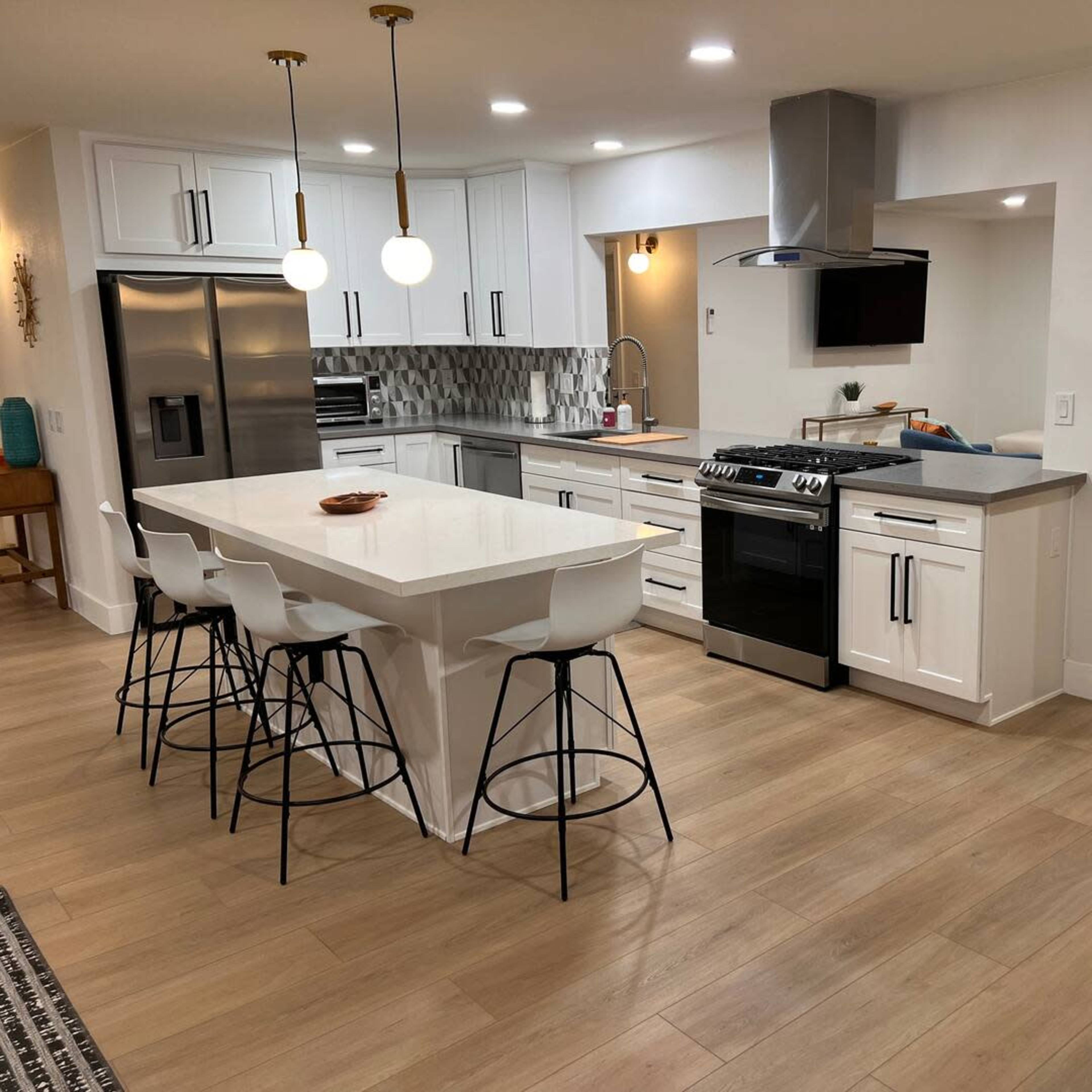 A modern kitchen with white cabinetry, a large island with seating, stainless steel appliances, and an adjoining living area.