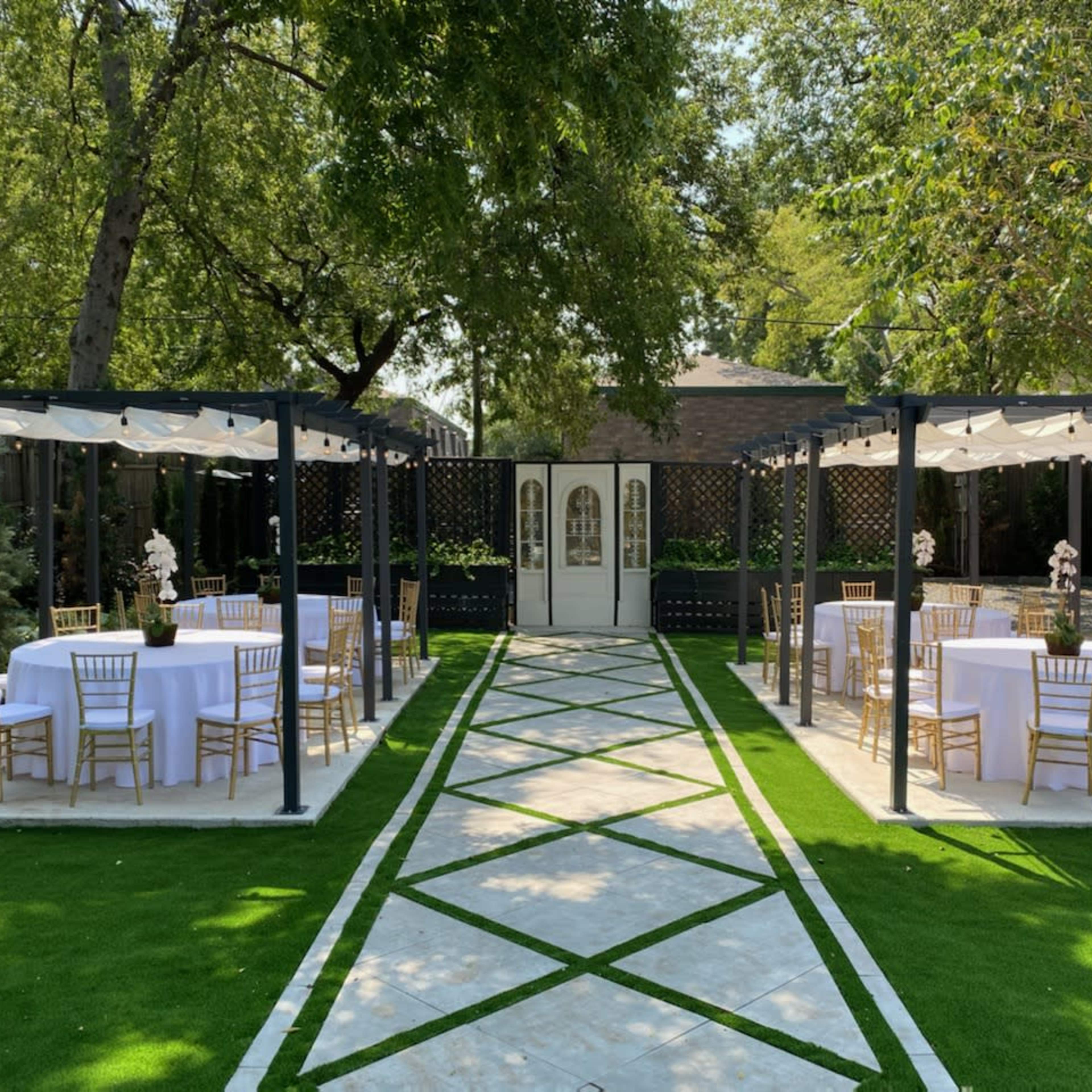 A landscaped outdoor area with two sets of dining tables and chairs arranged under pergolas, creating a symmetrical pathway leading to a set of double doors.