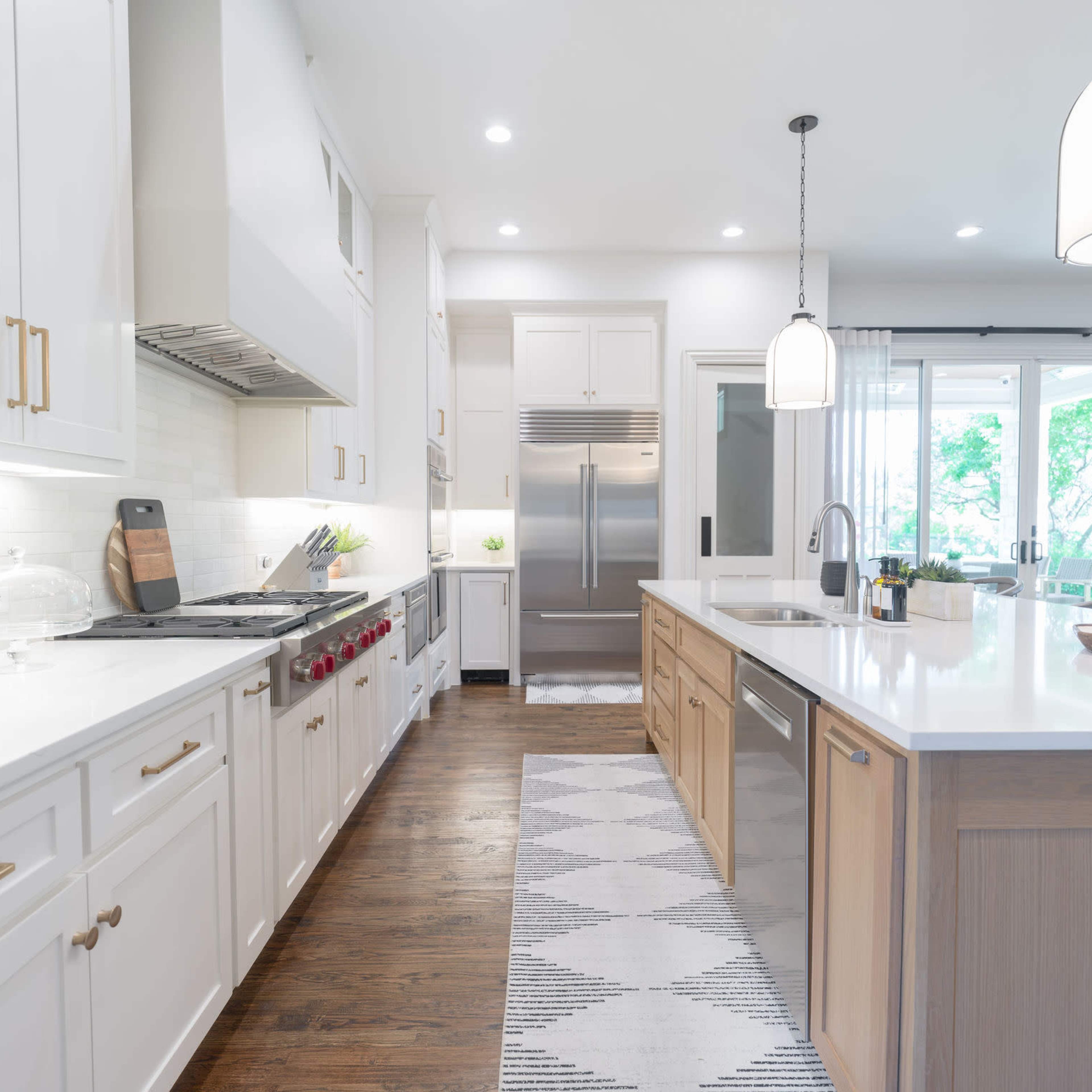 A modern kitchen featuring white cabinetry, stainless steel appliances, a large island, and a contemporary light fixture, with large windows providing natural light.