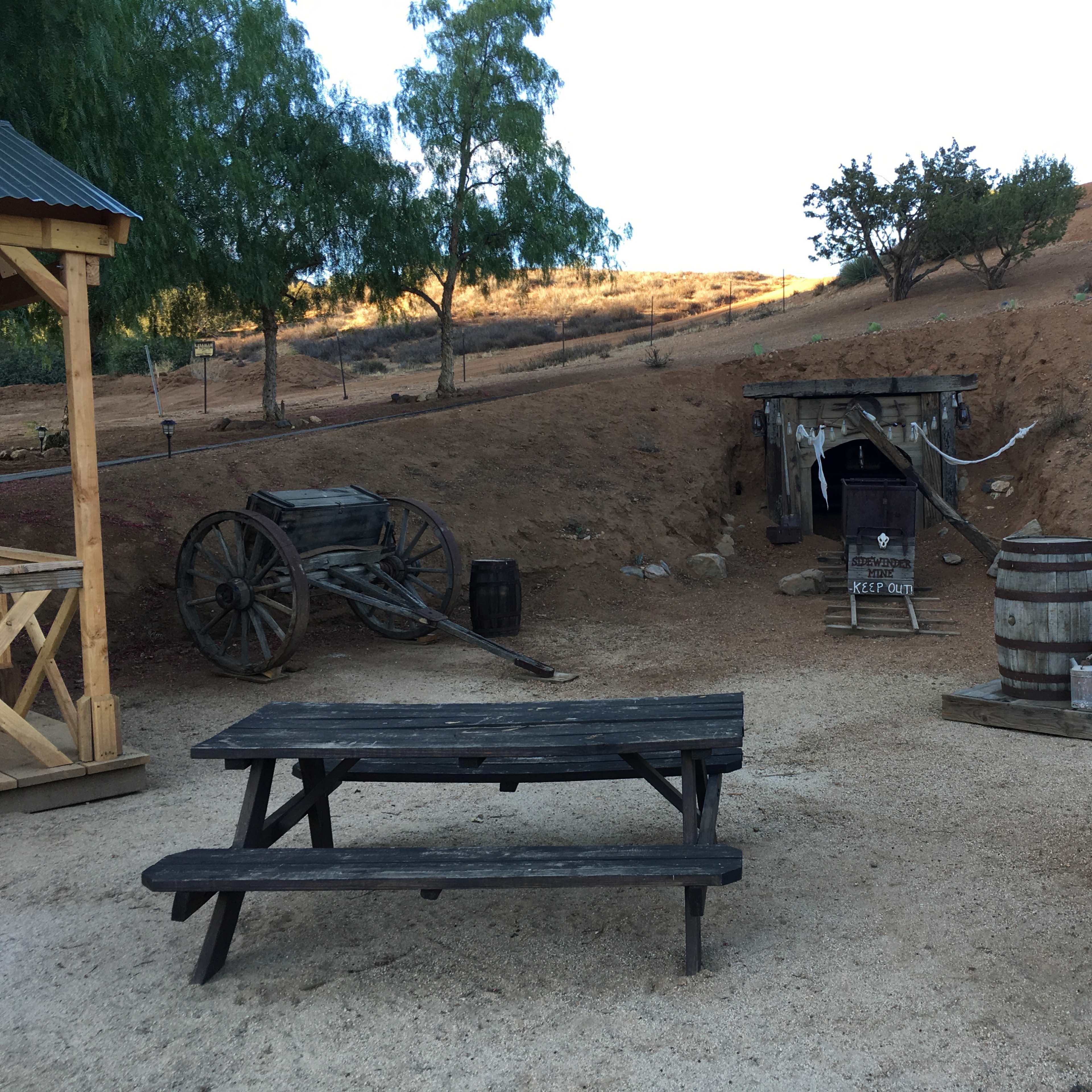 A rustic outdoor setting with a wooden wagon, a barrel, a well, and an area featuring a picnic table under a covered structure.