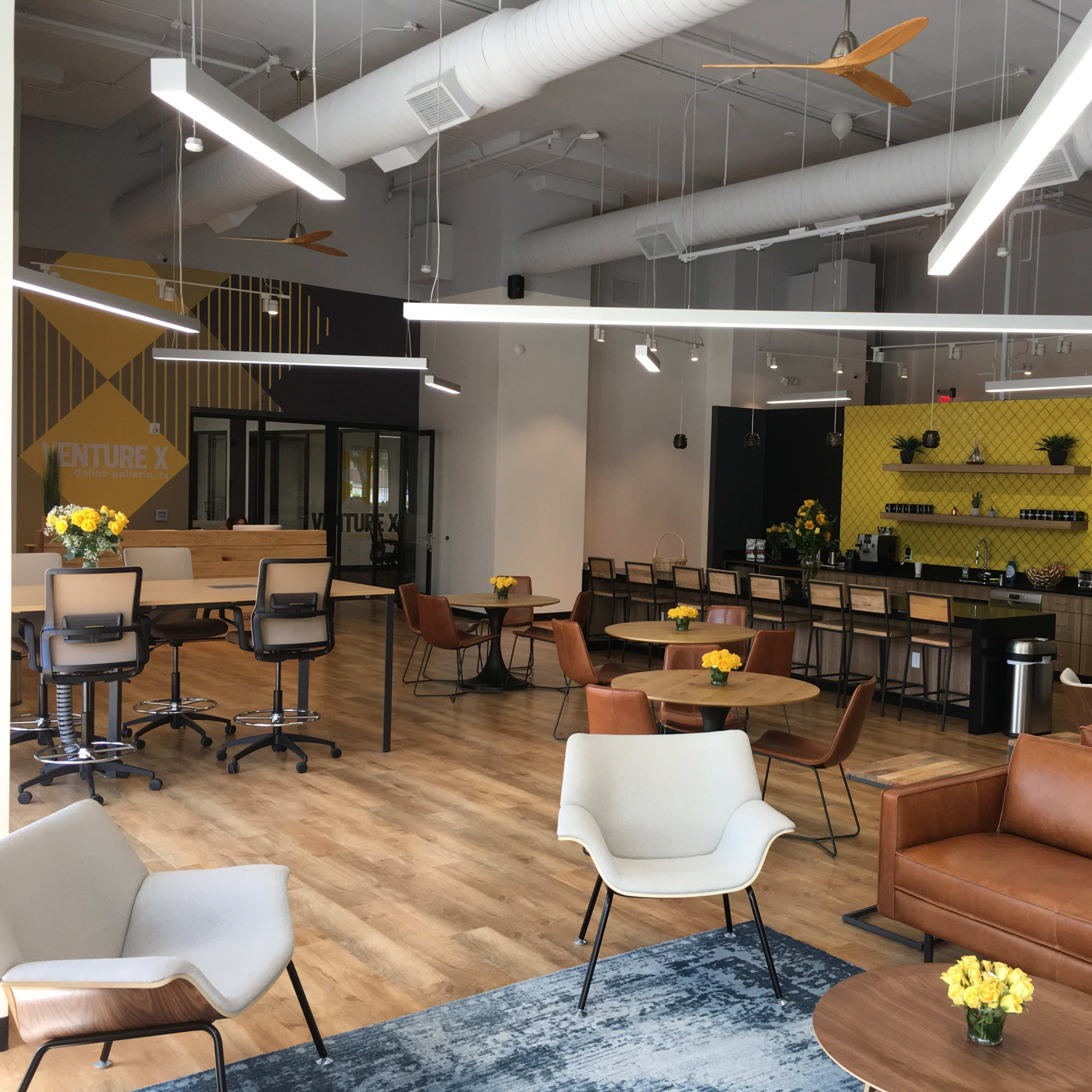 A modern coworking space with various seating areas, yellow flowers on tables, and a kitchenette in the background.