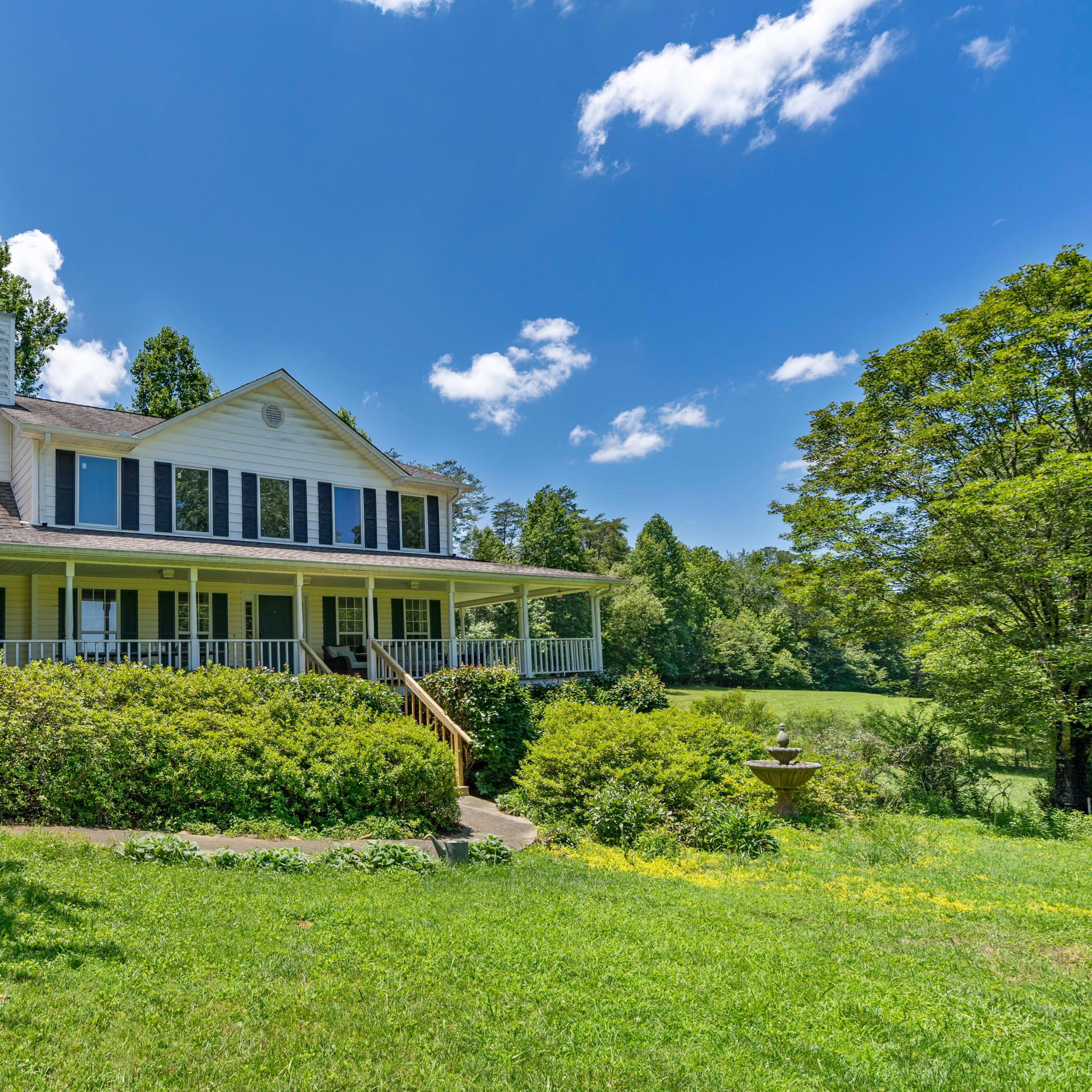 A large white house with a porch and blue shutters is surrounded by greenery under a clear blue sky.