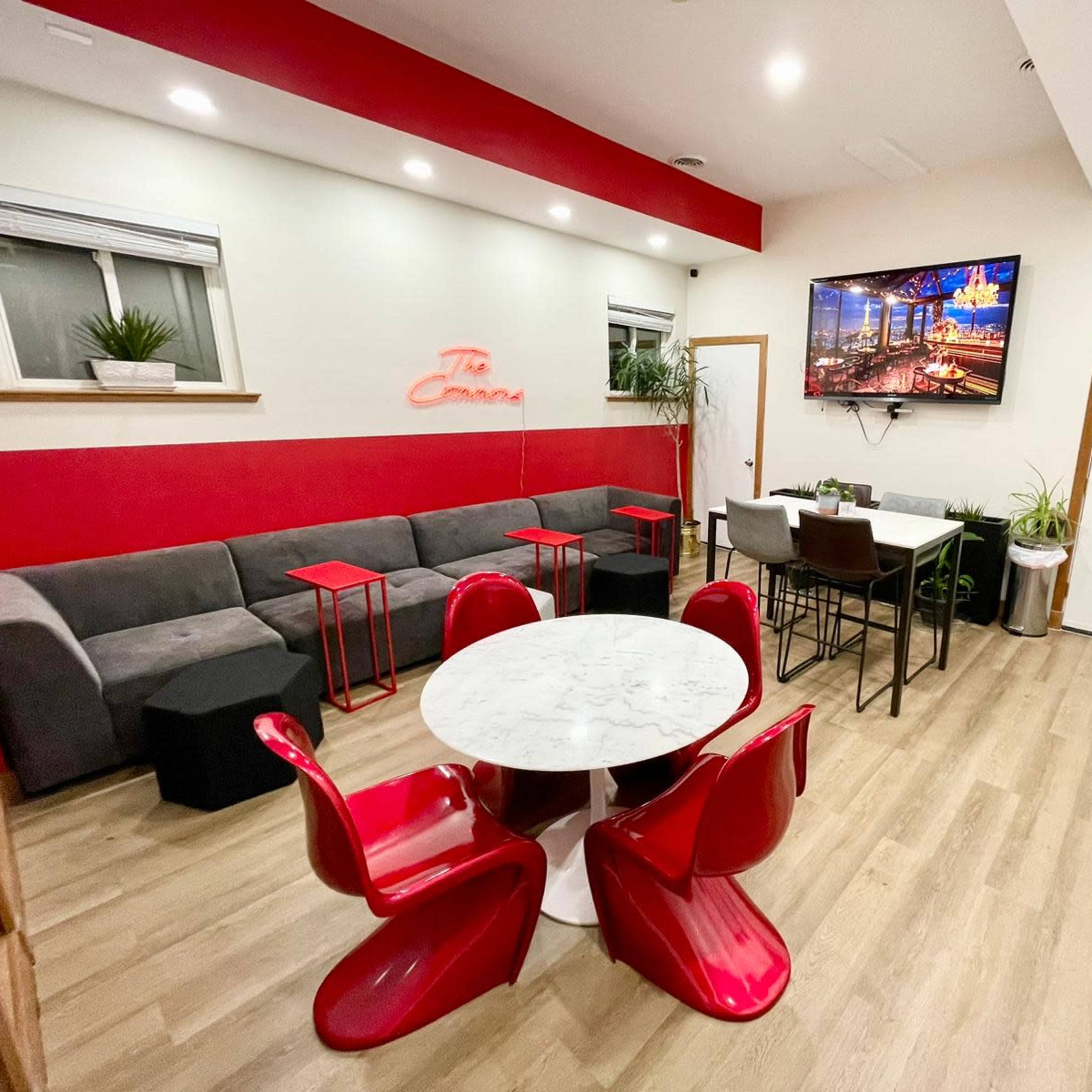 A modern, minimalist lounge area featuring a gray sofa, red accent walls, a marble table surrounded by red chairs, and a large TV mounted on the wall.