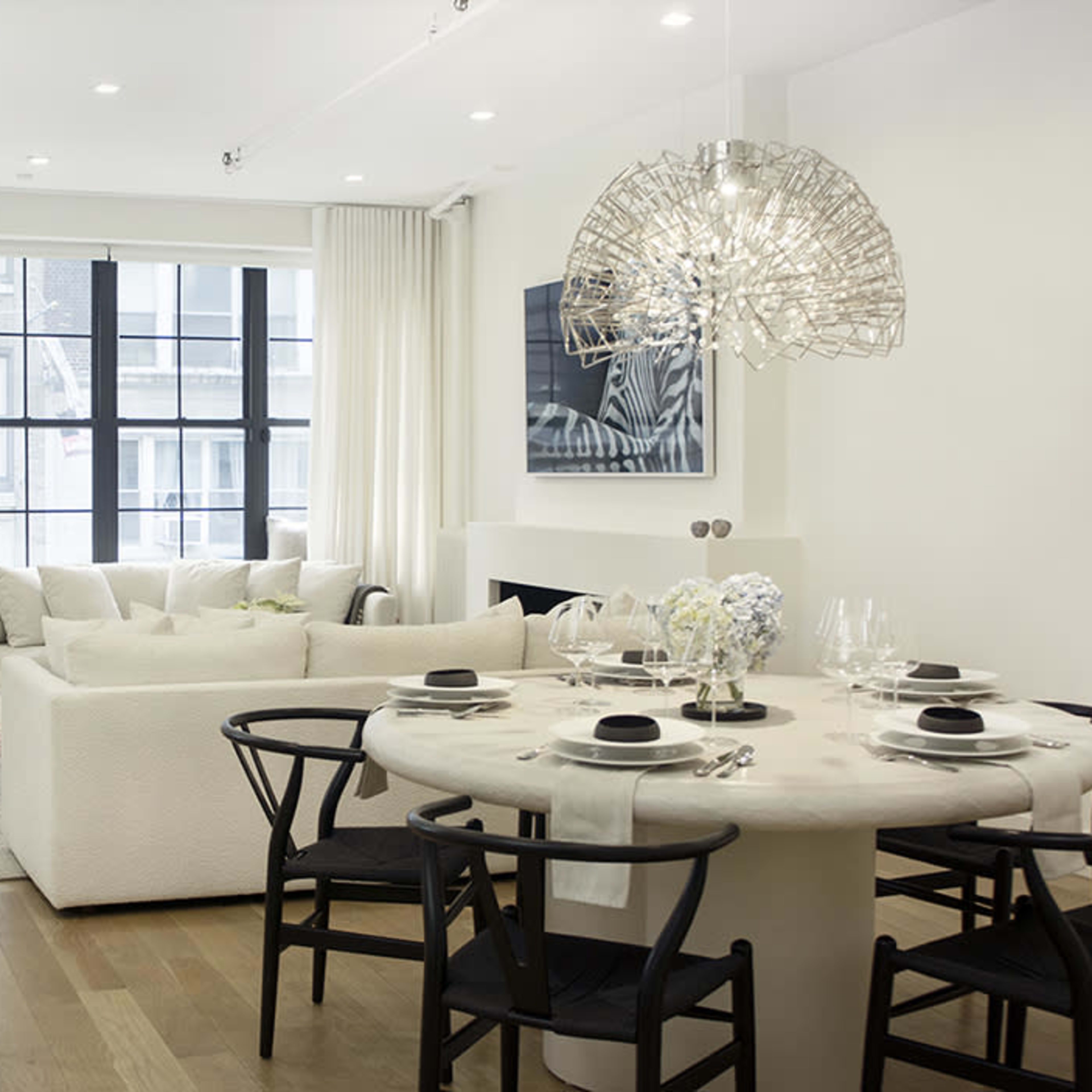 A modern interior living space featuring a round dining table with black chairs, a beige sofa, and large windows with sheer curtains.