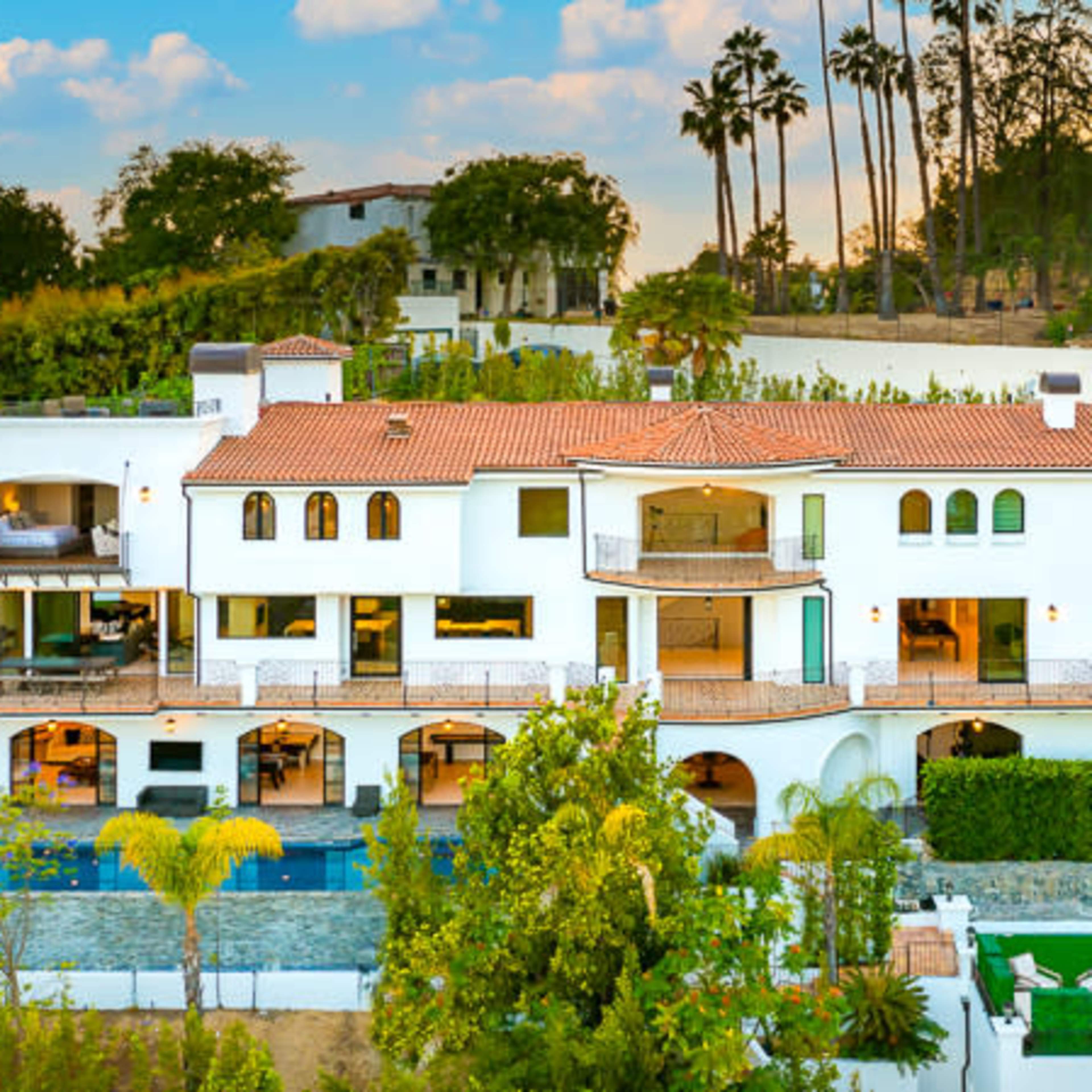 A large, white stucco villa with red tile roofing is situated on a hillside, surrounded by palm trees and lush greenery.