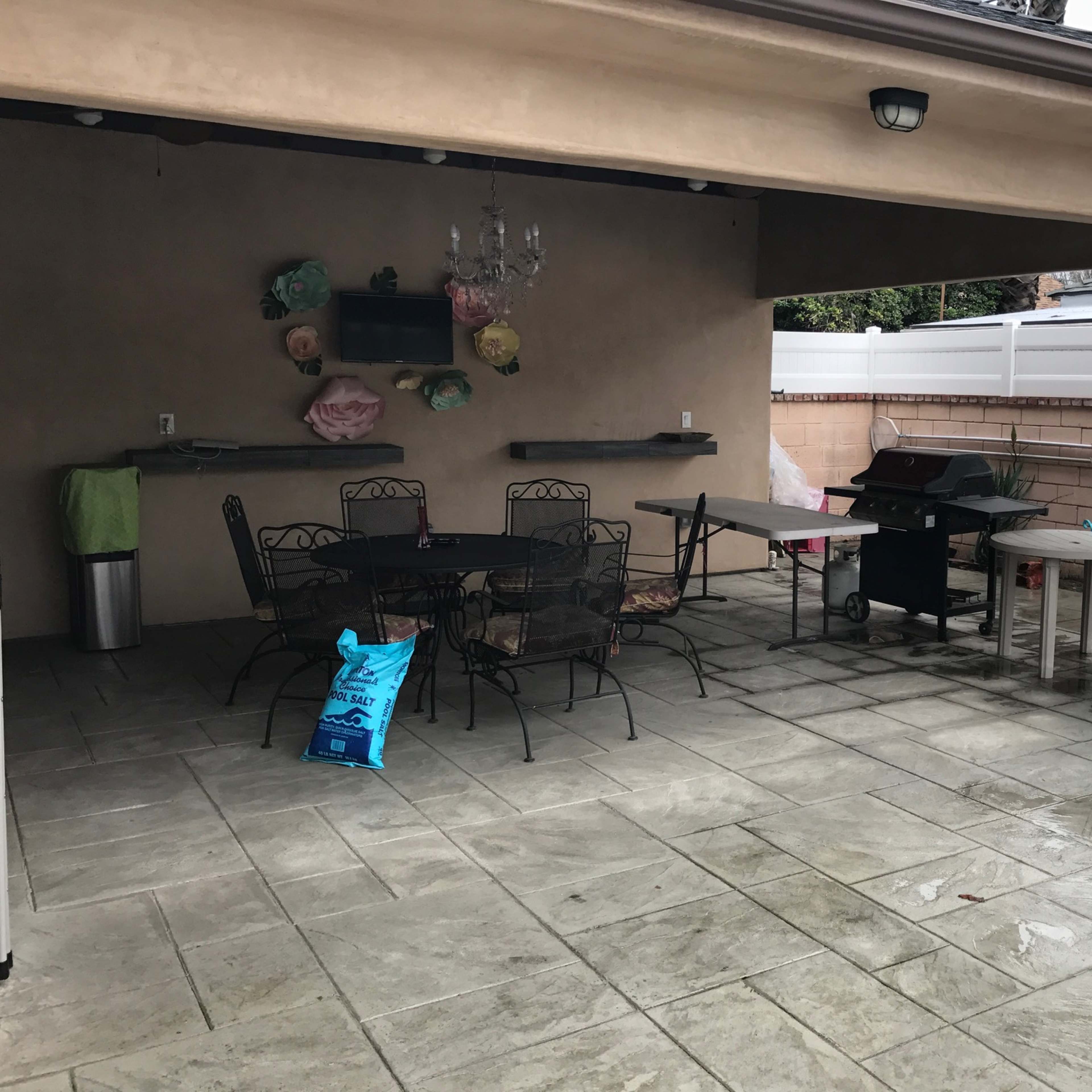 A covered patio features a dining set, a grill, and a small bistro table, with wet flooring from recent rain.