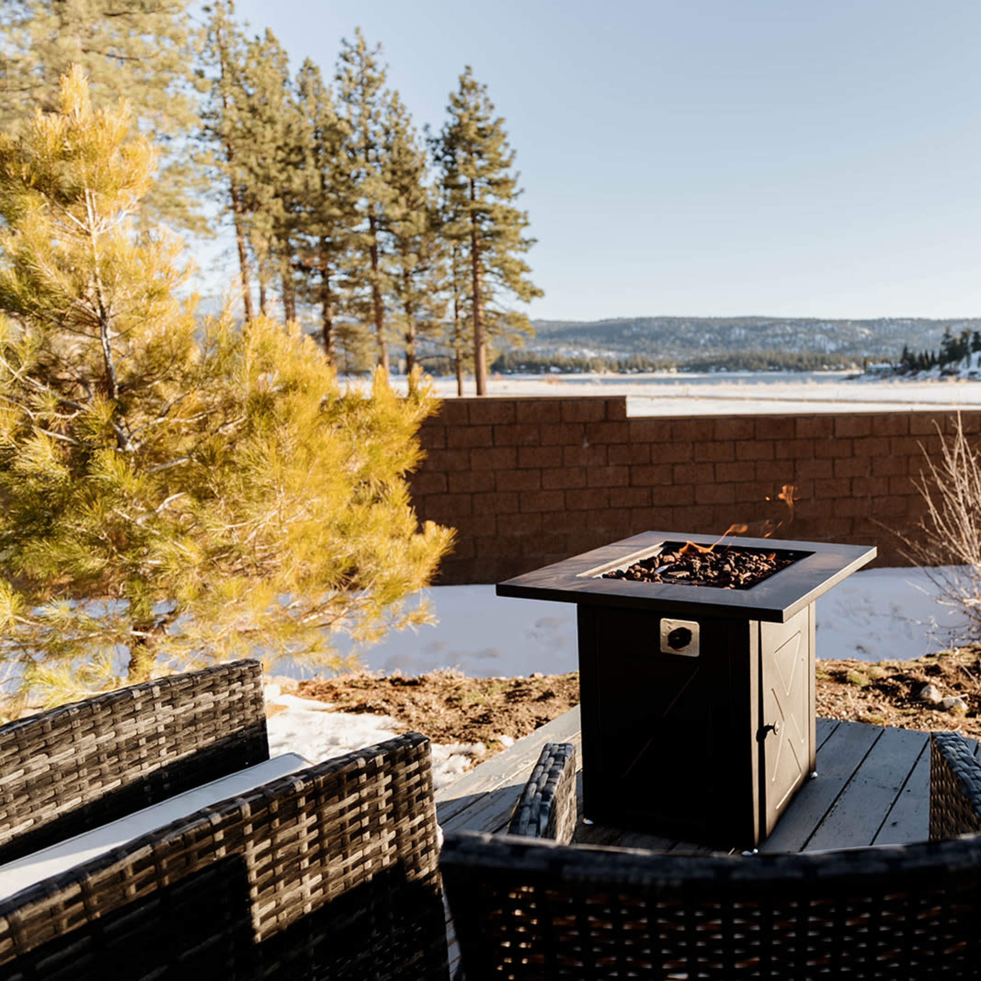 A fire pit surrounded by wicker chairs sits on a patio with a snow-covered landscape and tall pine trees in the background.