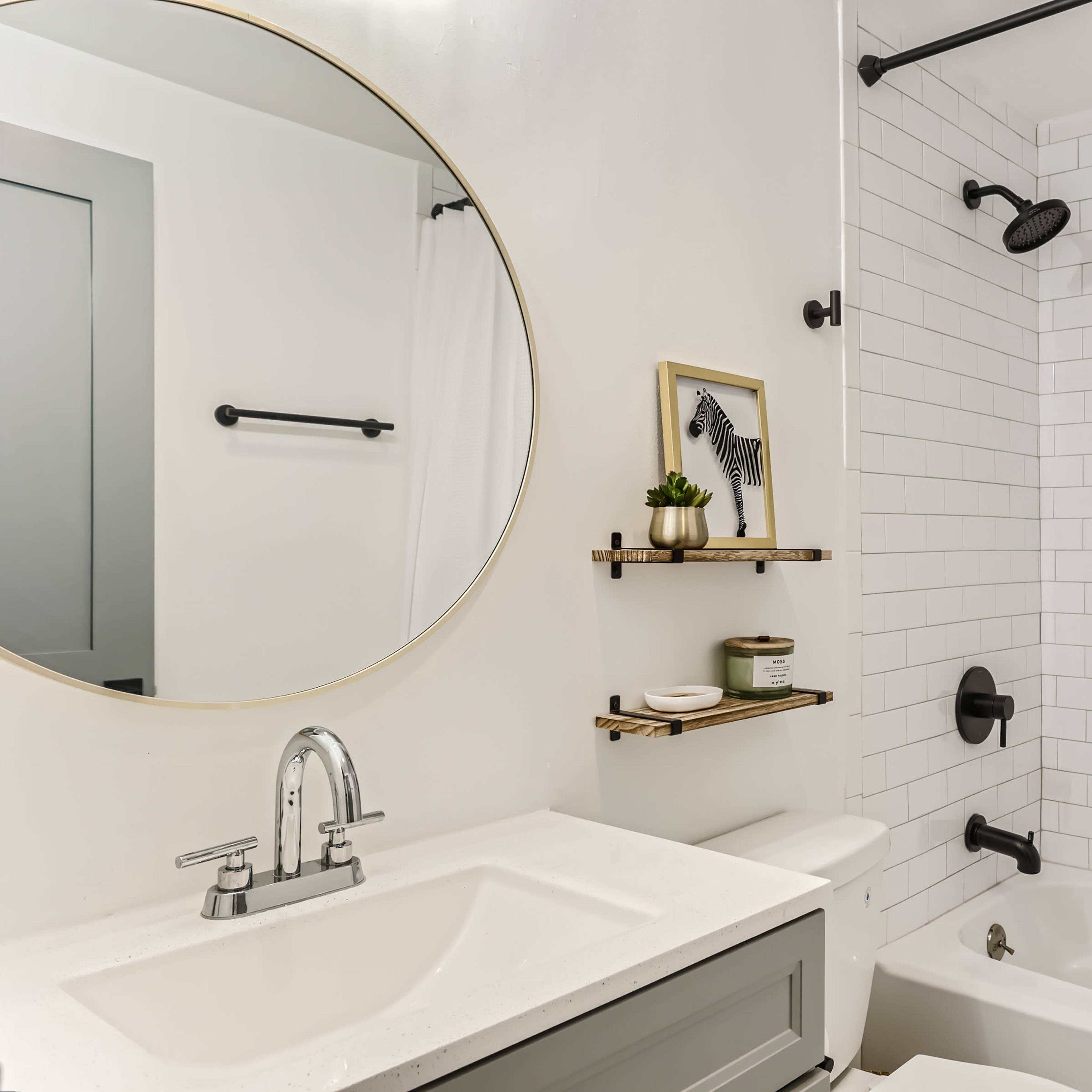 A modern bathroom with a round mirror above a sink, a shower with a black showerhead, and white tiled walls featuring a decorative inset.
