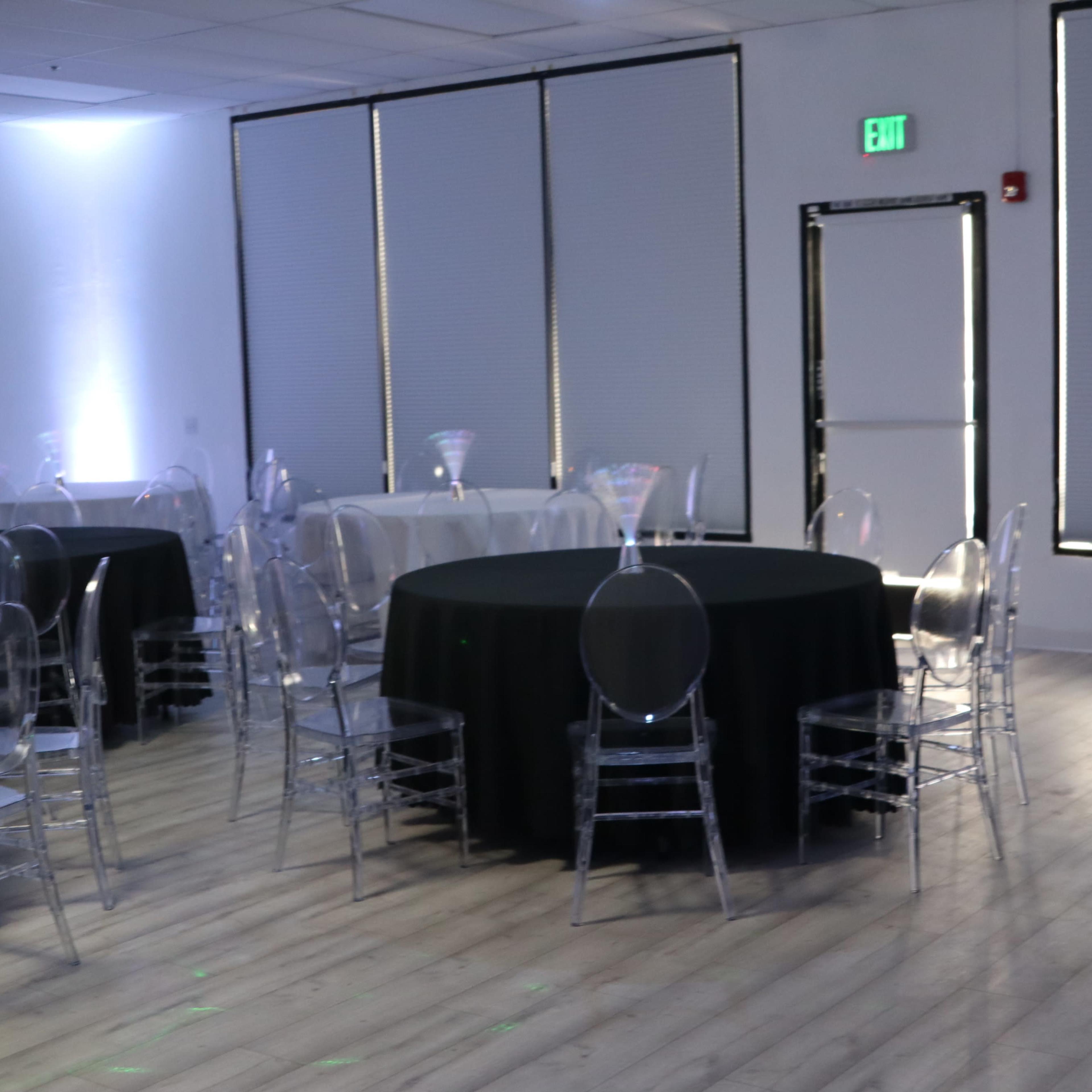 A spacious event room with several round tables covered in black and white linens, surrounded by clear acrylic chairs, and illuminated by soft lighting.