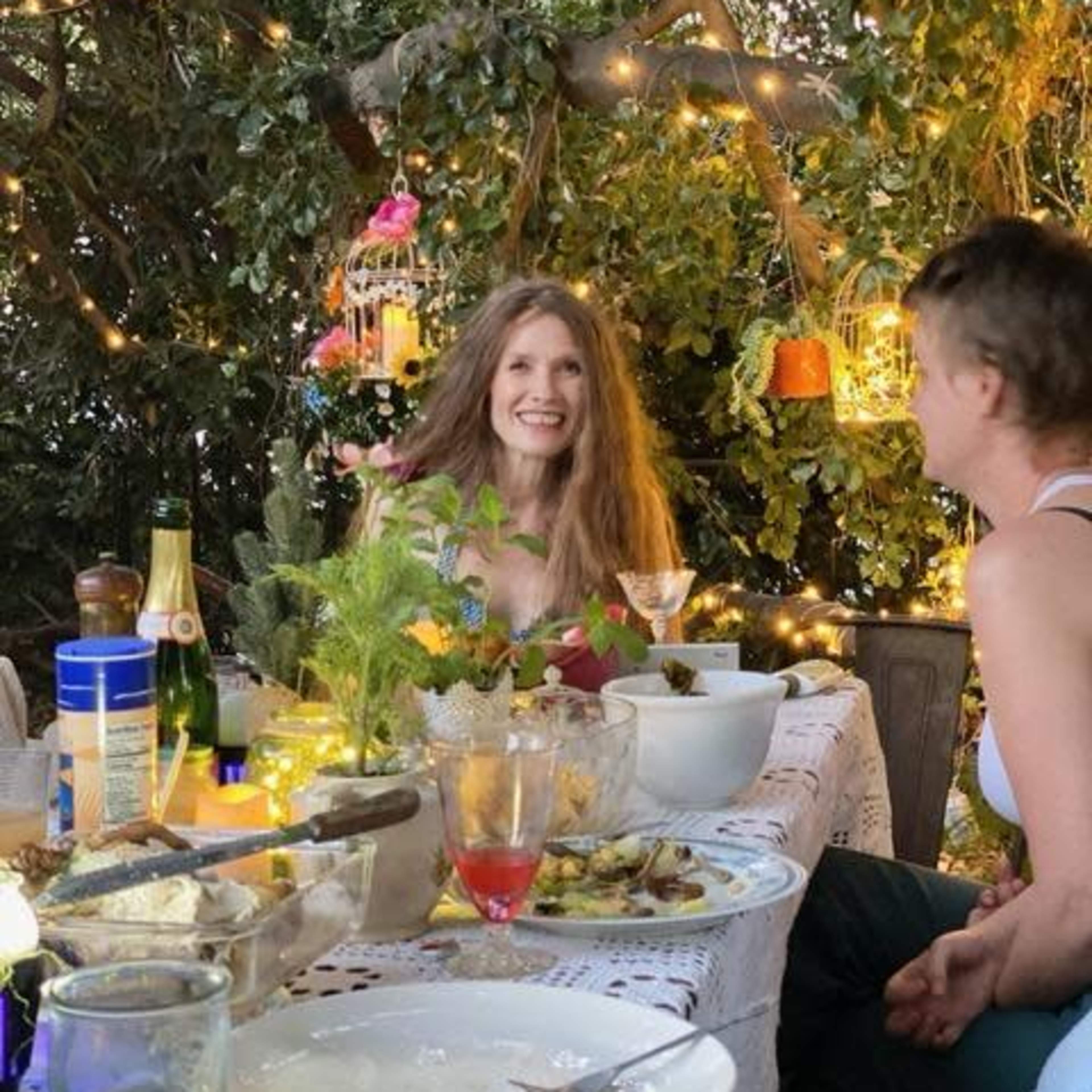 A woman with long hair sits at a table adorned with food and drinks, surrounded by greenery and decorative lights.