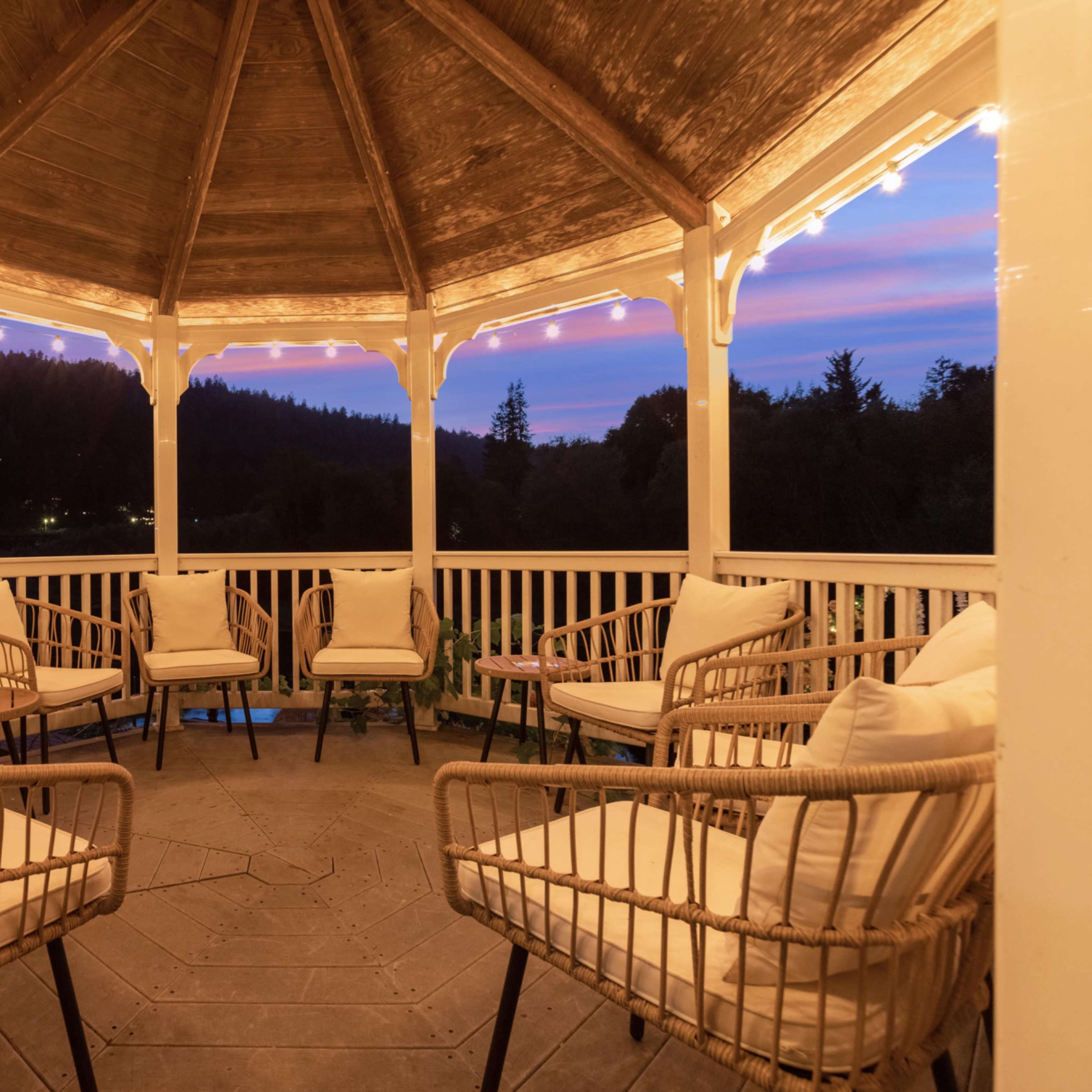 A gazebo with wicker chairs is illuminated by string lights against a sunset backdrop of mountains and sky.