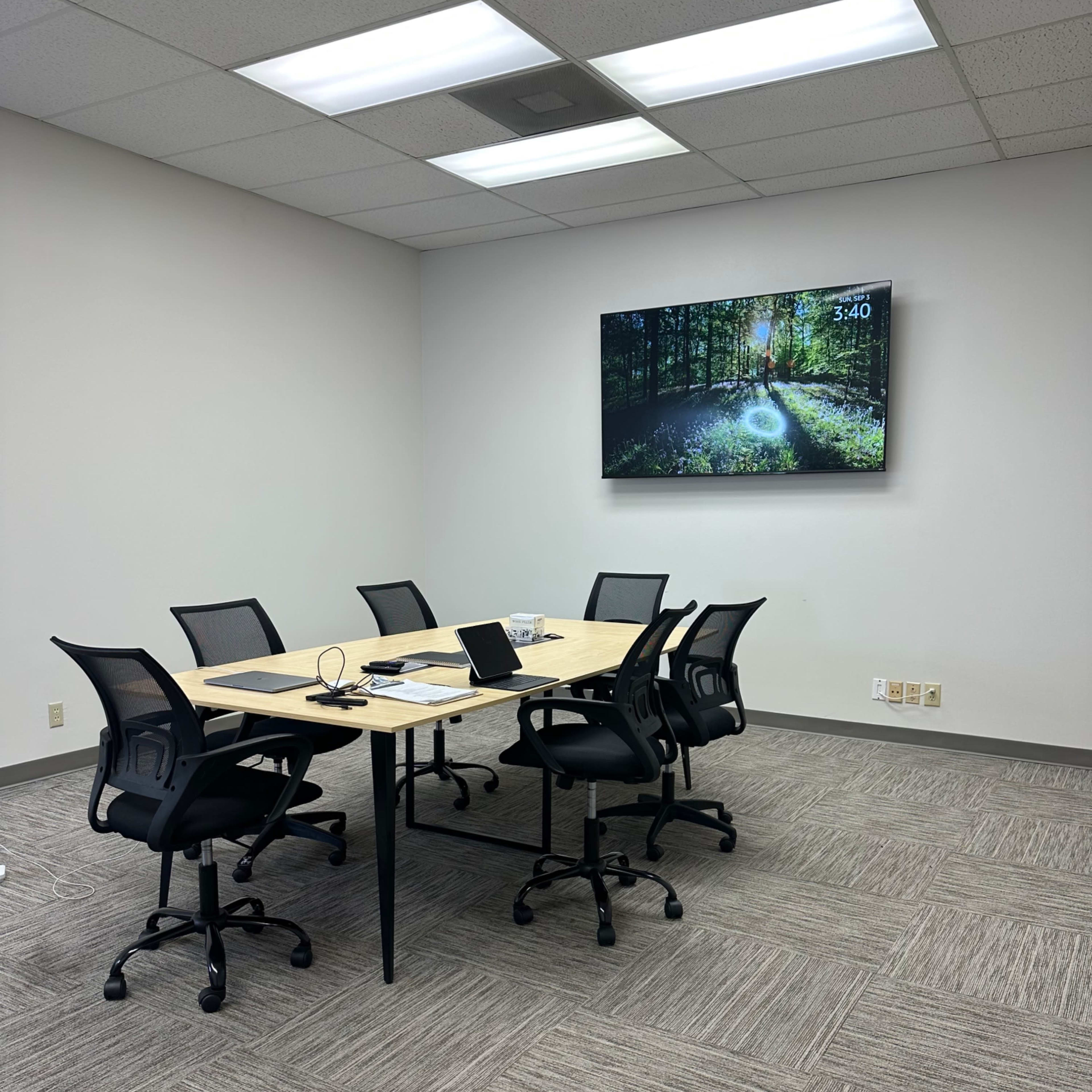 A modern conference room features a rectangular table with six black chairs, a laptop, and a television displaying a forest scene on the wall.