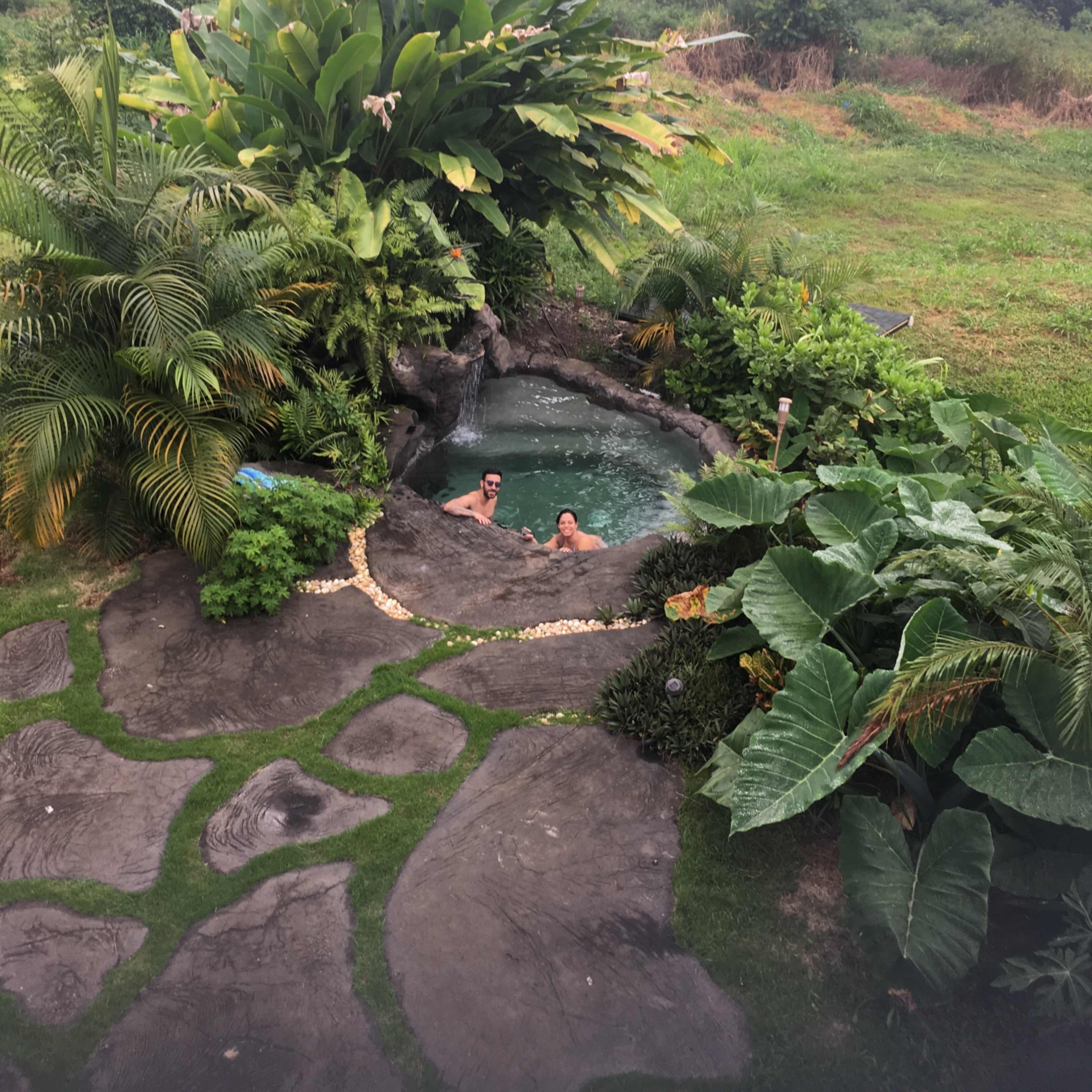 Two people relax in a hot tub surrounded by lush greenery and stone pathways in a landscaped garden.
