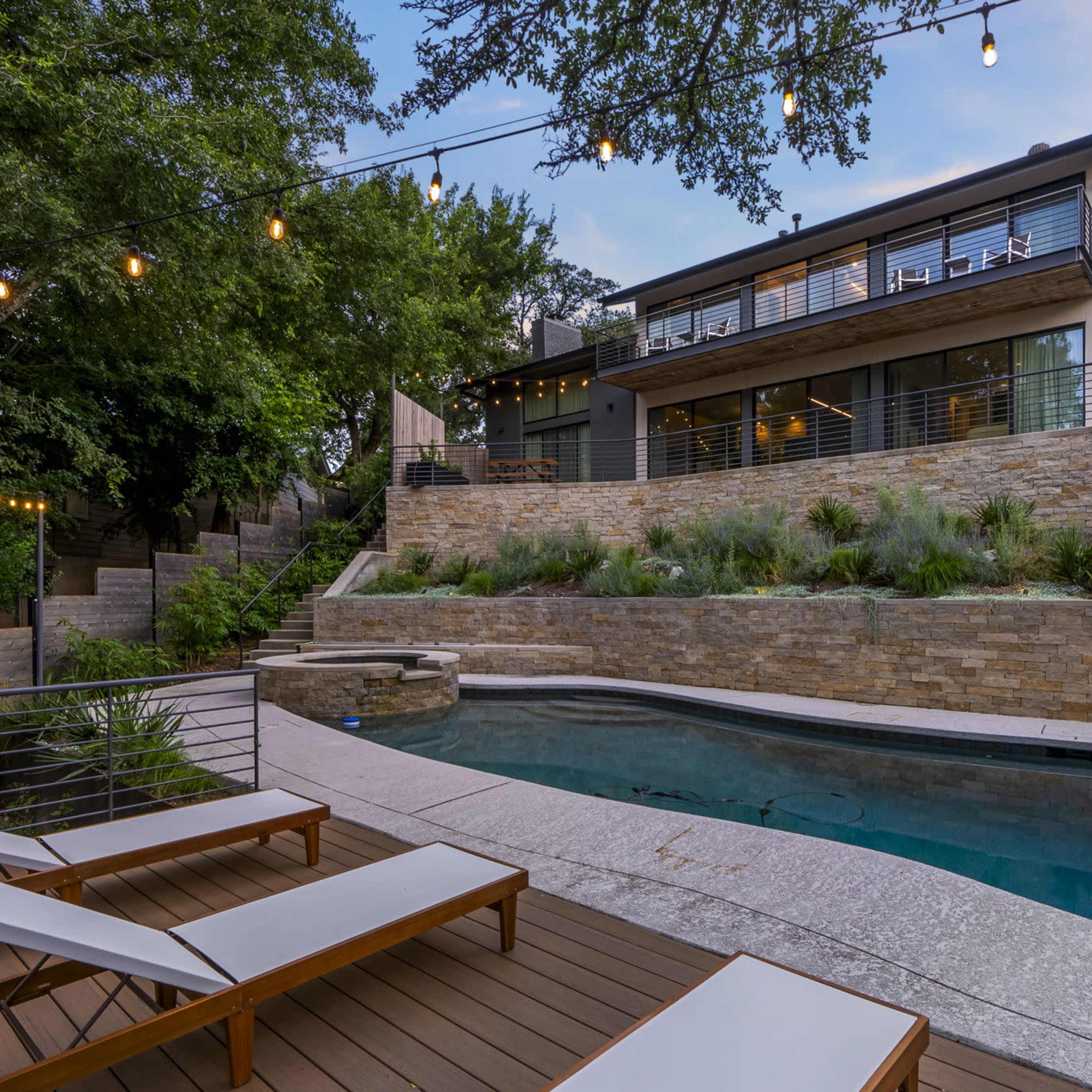A modern home with large windows overlooking a landscaped yard and a swimming pool, framed by string lights and wooden loungers.